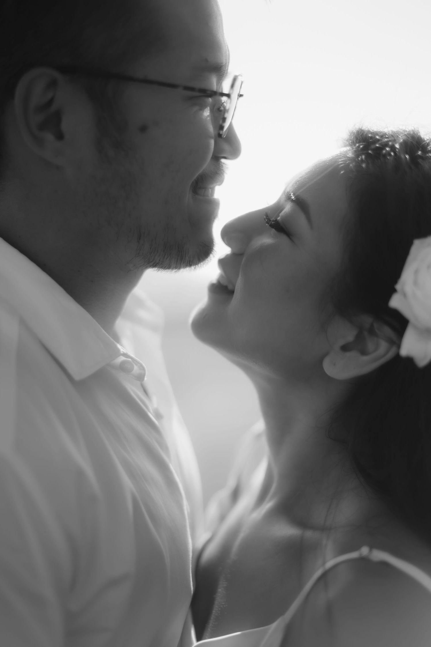 Black and white photo of a smiling couple gazing at each other intimately, man wearing glasses and white shirt.