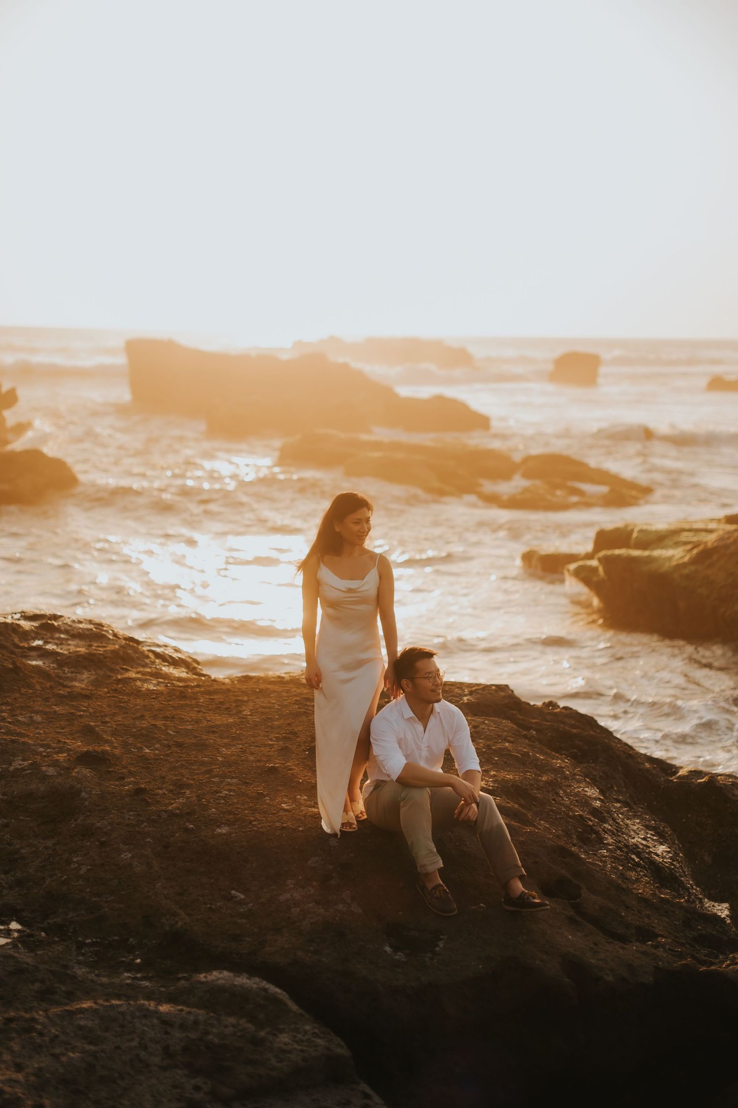 Couple in white attire posing on rocky coastal cliffs at golden sunset with ocean waves in background.