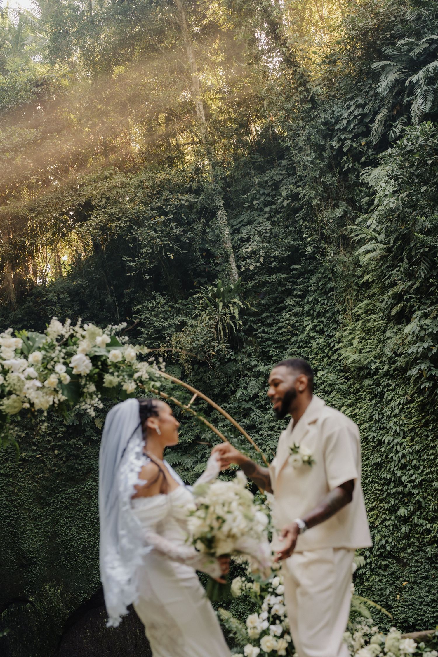 Bride and groom dance beneath a golden floral arch with sunbeams in a lush green jungle.