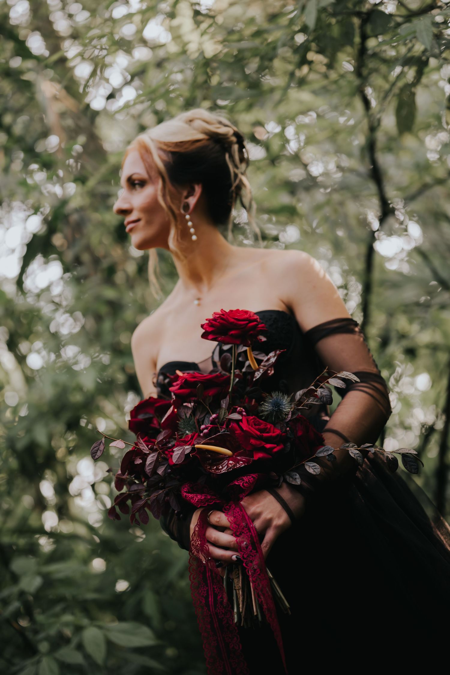 A black-gowned figure holds a dramatic bouquet of deep red roses and dark foliage in a moody outdoor setting.