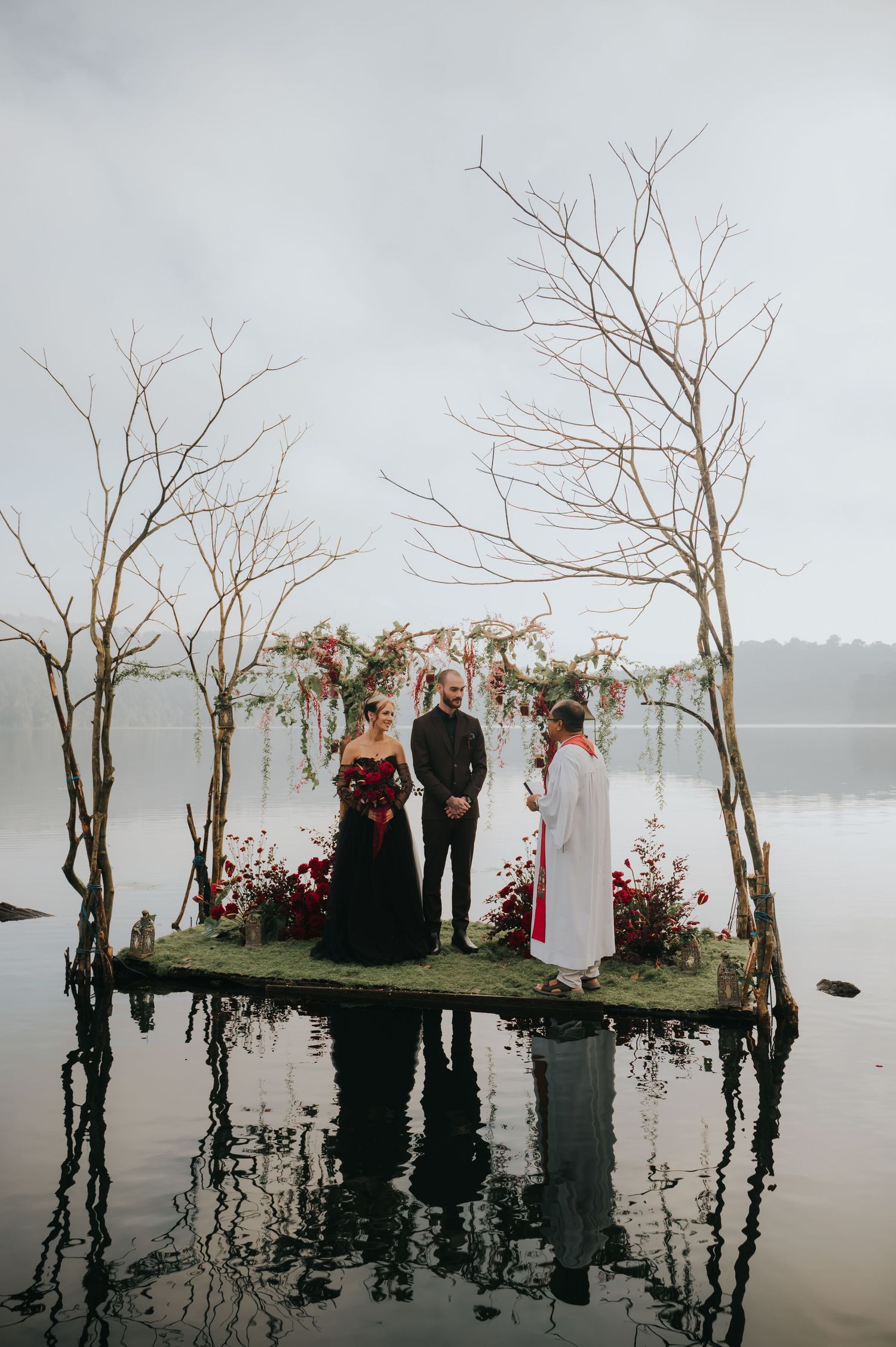 A wedding ceremony takes place on a floating platform over a misty lake with bare trees and floral decorations.