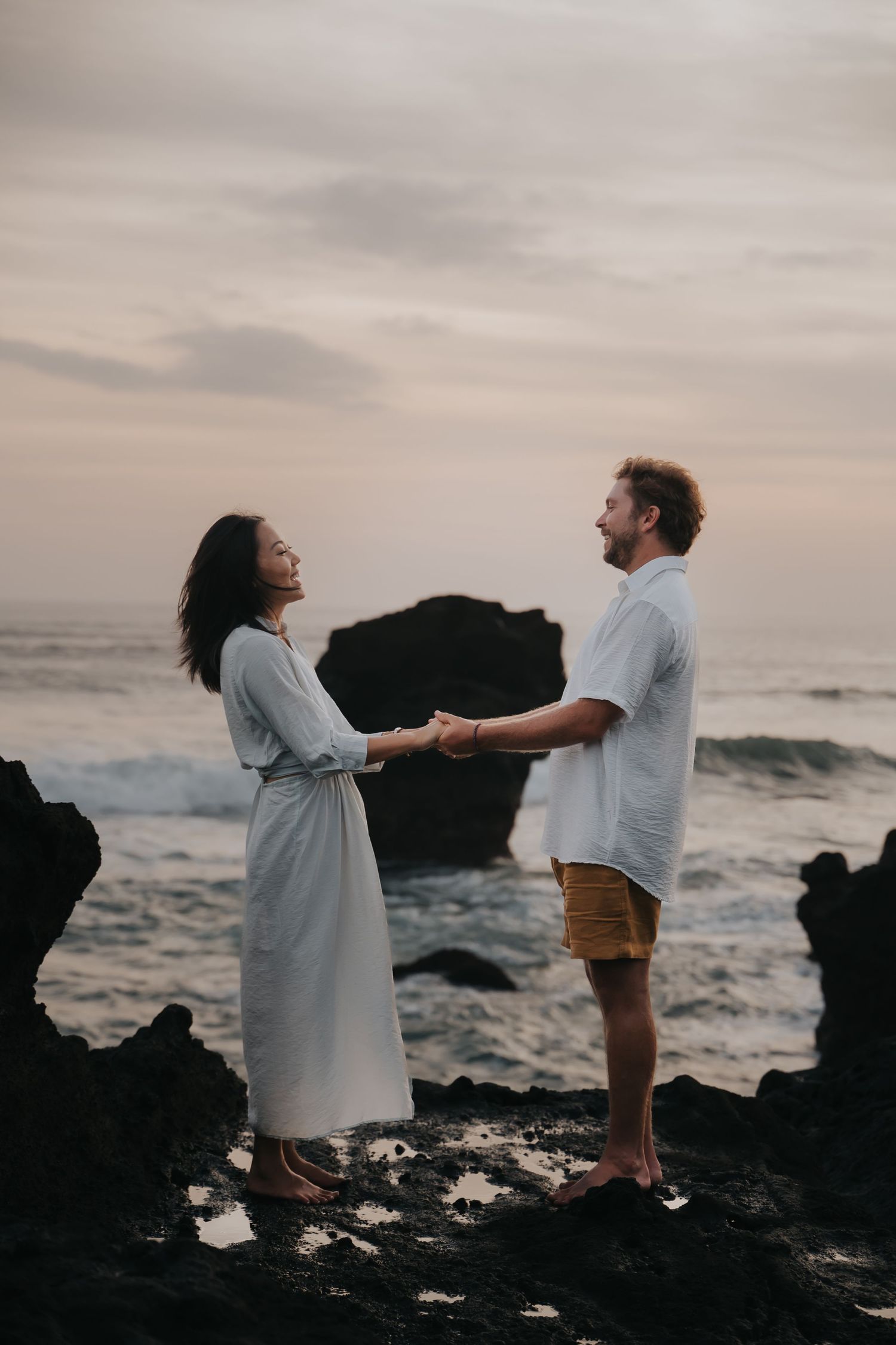 A couple dances together on rocky coastal cliffs at sunset with ocean waves crashing in the background.