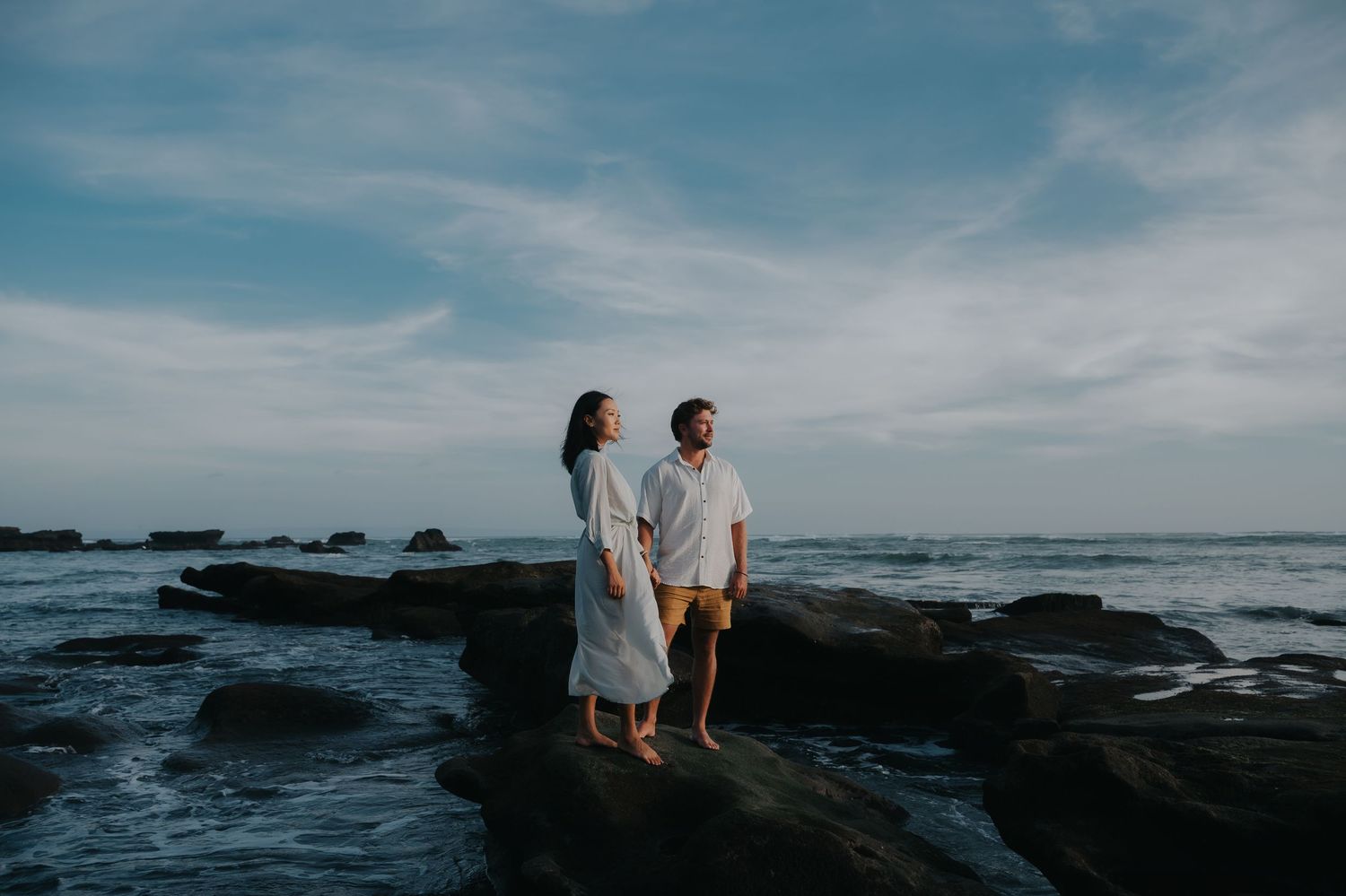 A couple walks along a rocky coastline at sunset with dramatic clouds and ocean waves in the background.
