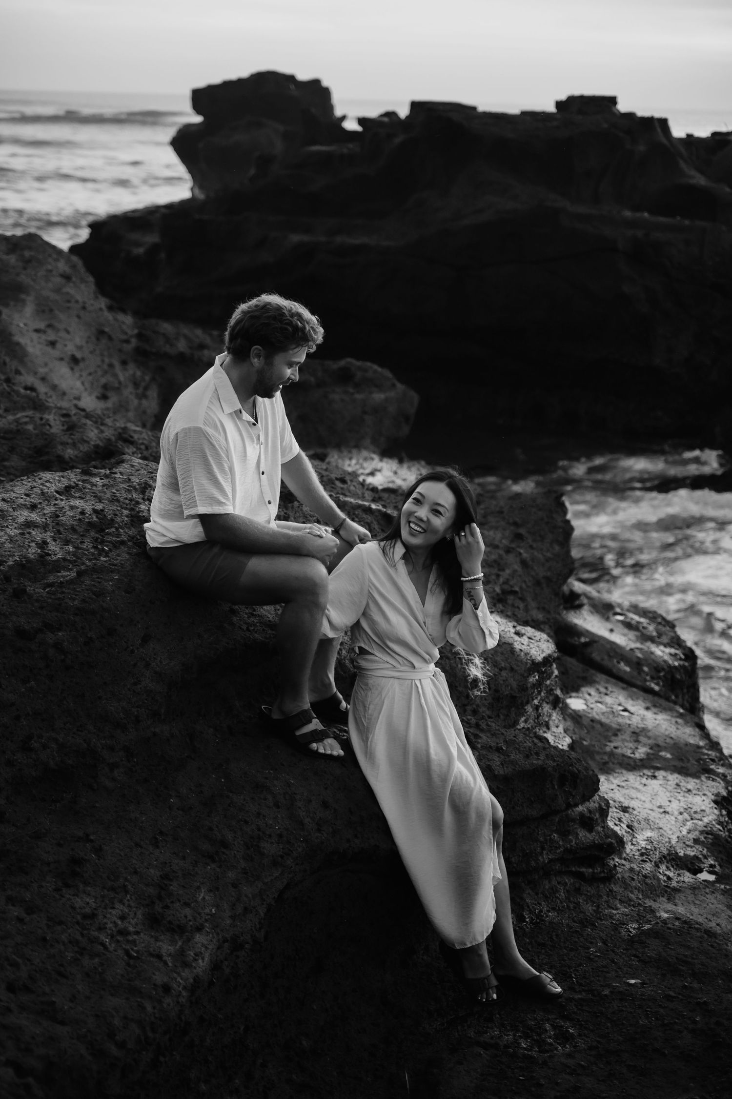 A couple shares a joyful moment together while sitting on coastal rocks near the ocean in black and white photography.