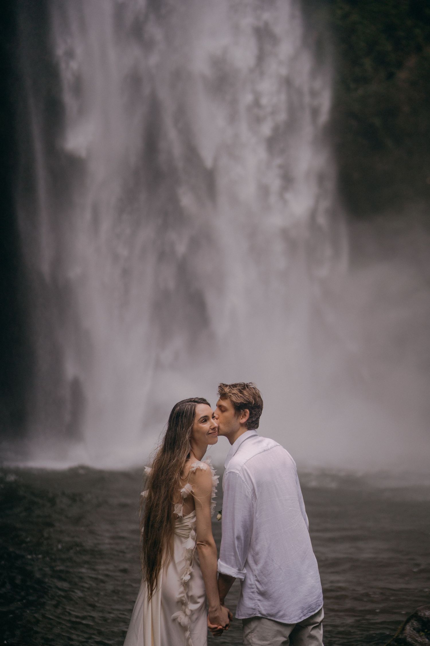 A romantic couple embraces near a misty waterfall in a dreamy outdoor photoshoot.