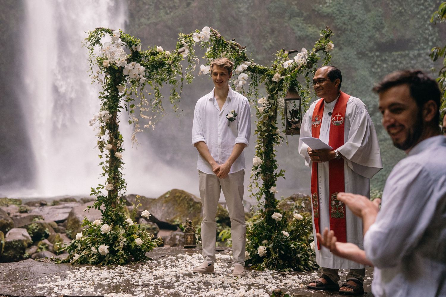 A group of people stand together in front of a white wall adorned with climbing vines and roses during a ceremony.