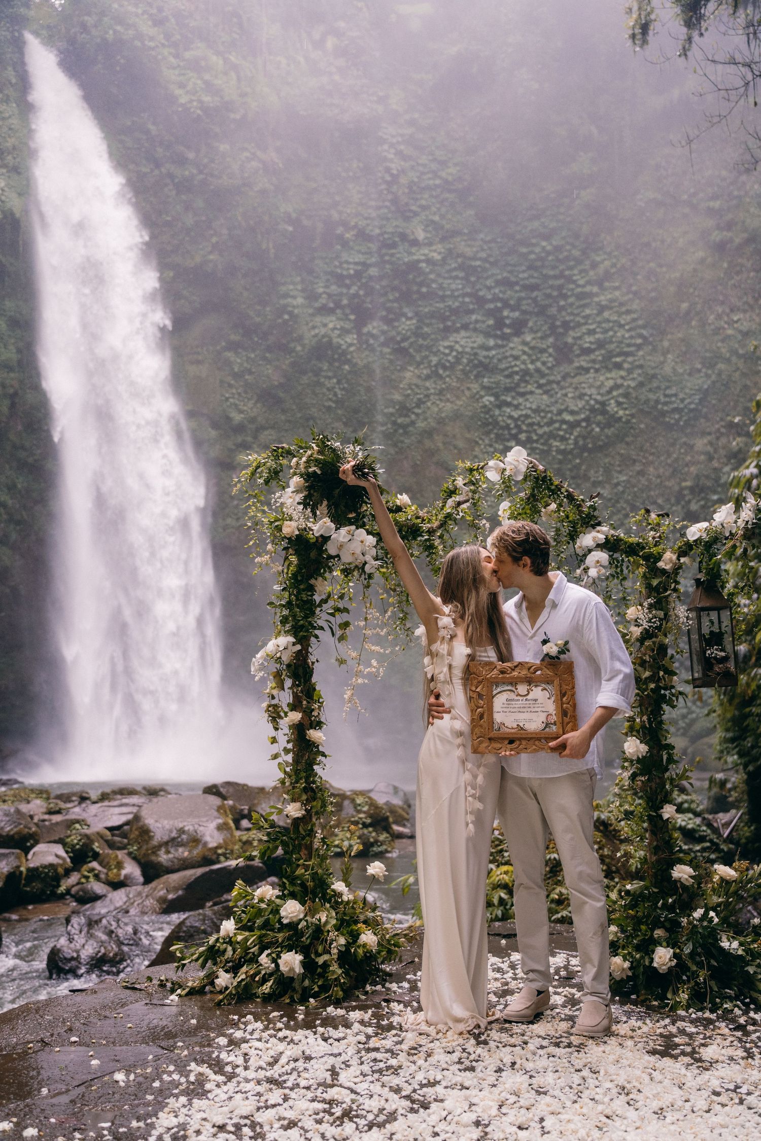 A romantic wedding ceremony takes place in front of a majestic waterfall with a floral arch adorned with white and green blooms.