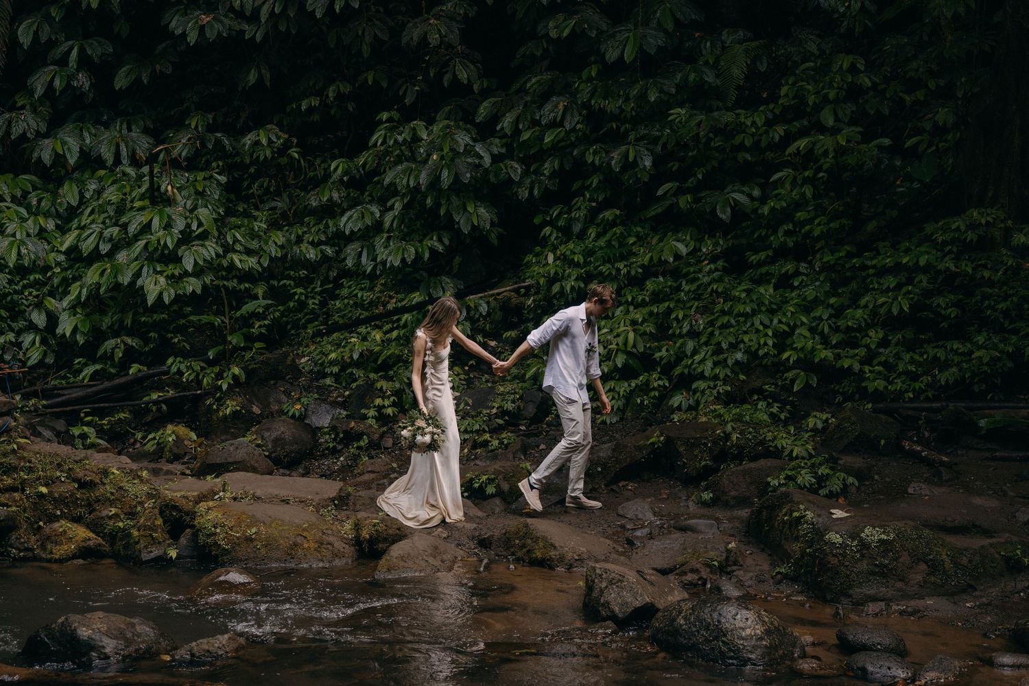 A couple walks hand in hand through a scenic wooded creek bed surrounded by lush greenery.