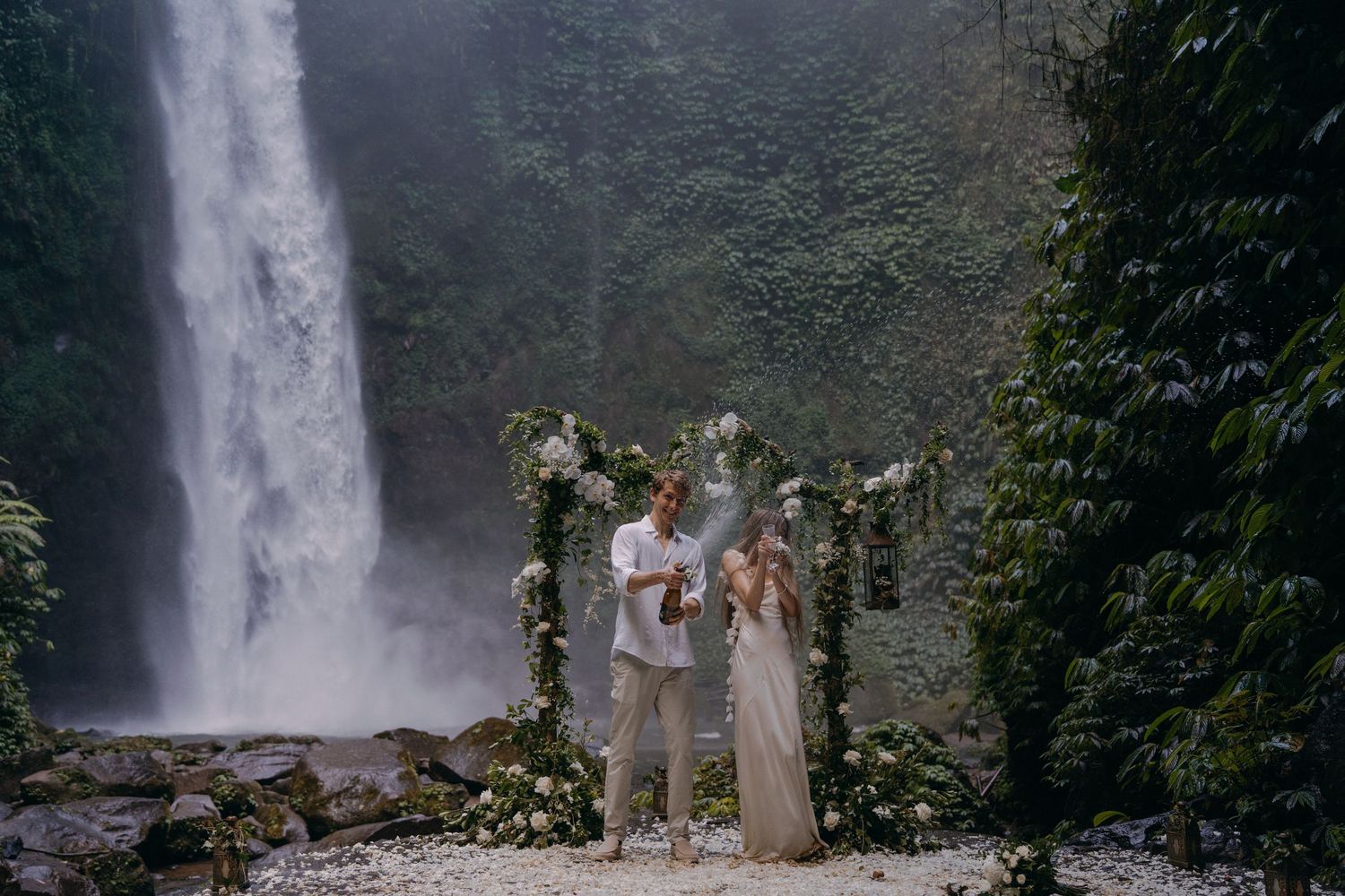 A group of people stand near a majestic waterfall in a lush forest setting, creating a scenic outdoor moment.