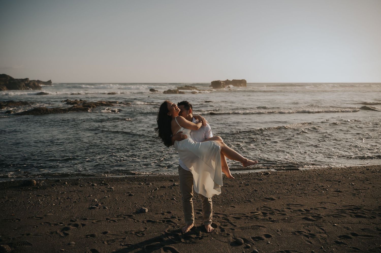 A couple shares a romantic embrace on a sandy beach at sunset, with ocean waves and rocky outcrops in the background.