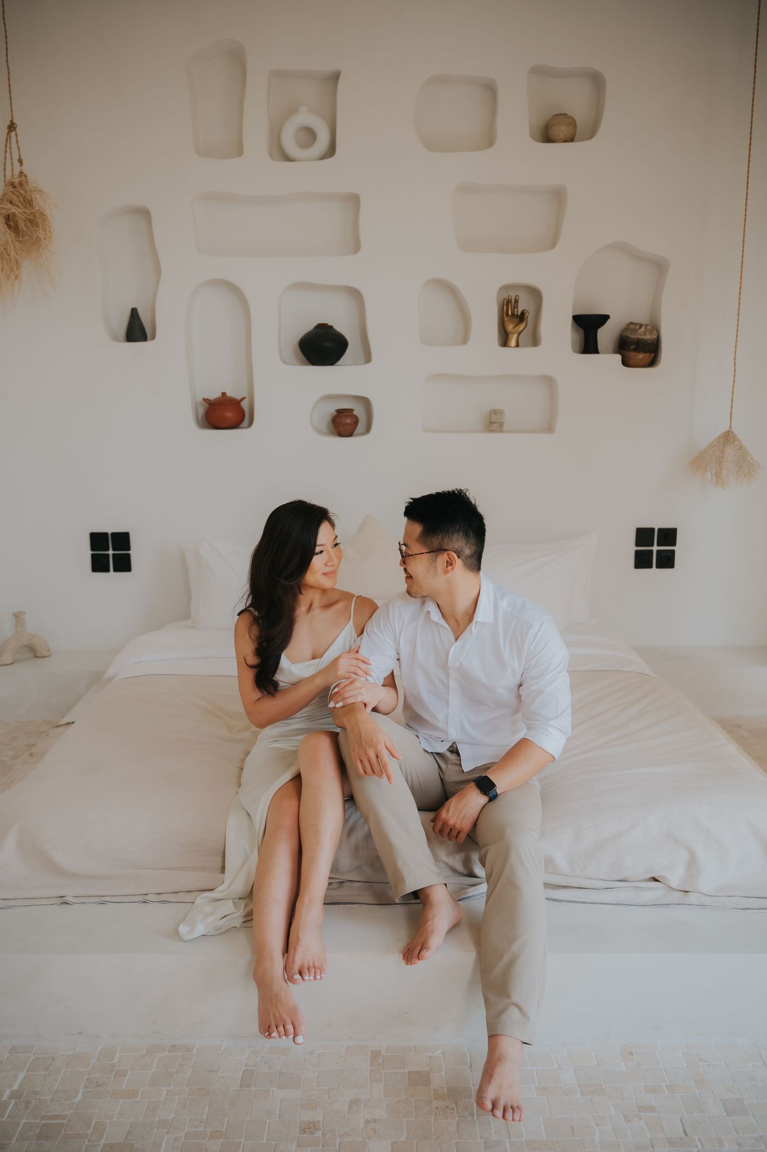 A couple sits barefoot on a white bed, smiling at each other in a bright boho-style room with decorative wall niches.