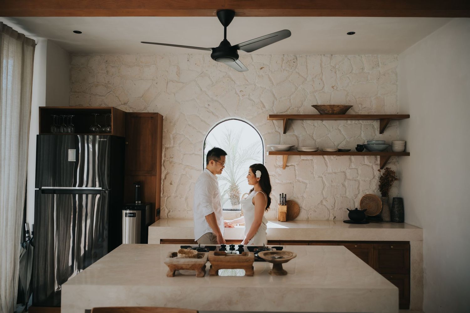 Couple sharing a romantic moment in a modern boho kitchen with arched window, open shelves, and a concrete island.
