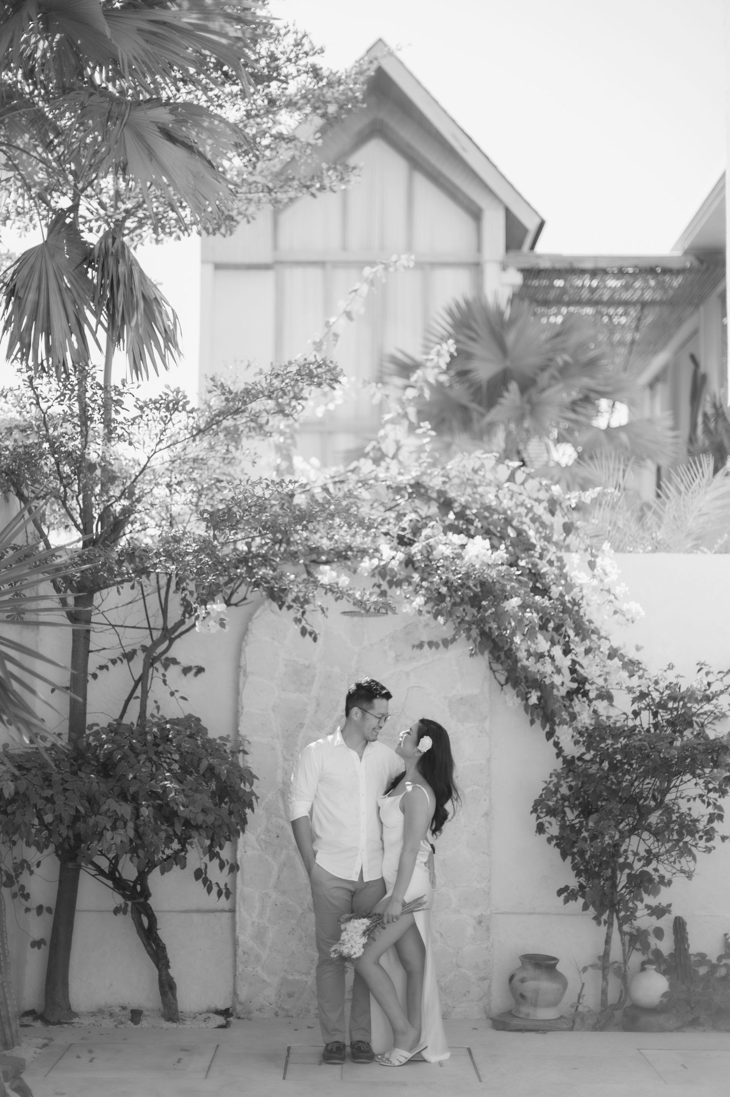 Black and white photo of a romantic couple embracing under a floral arch beside a white wall with tropical plants.