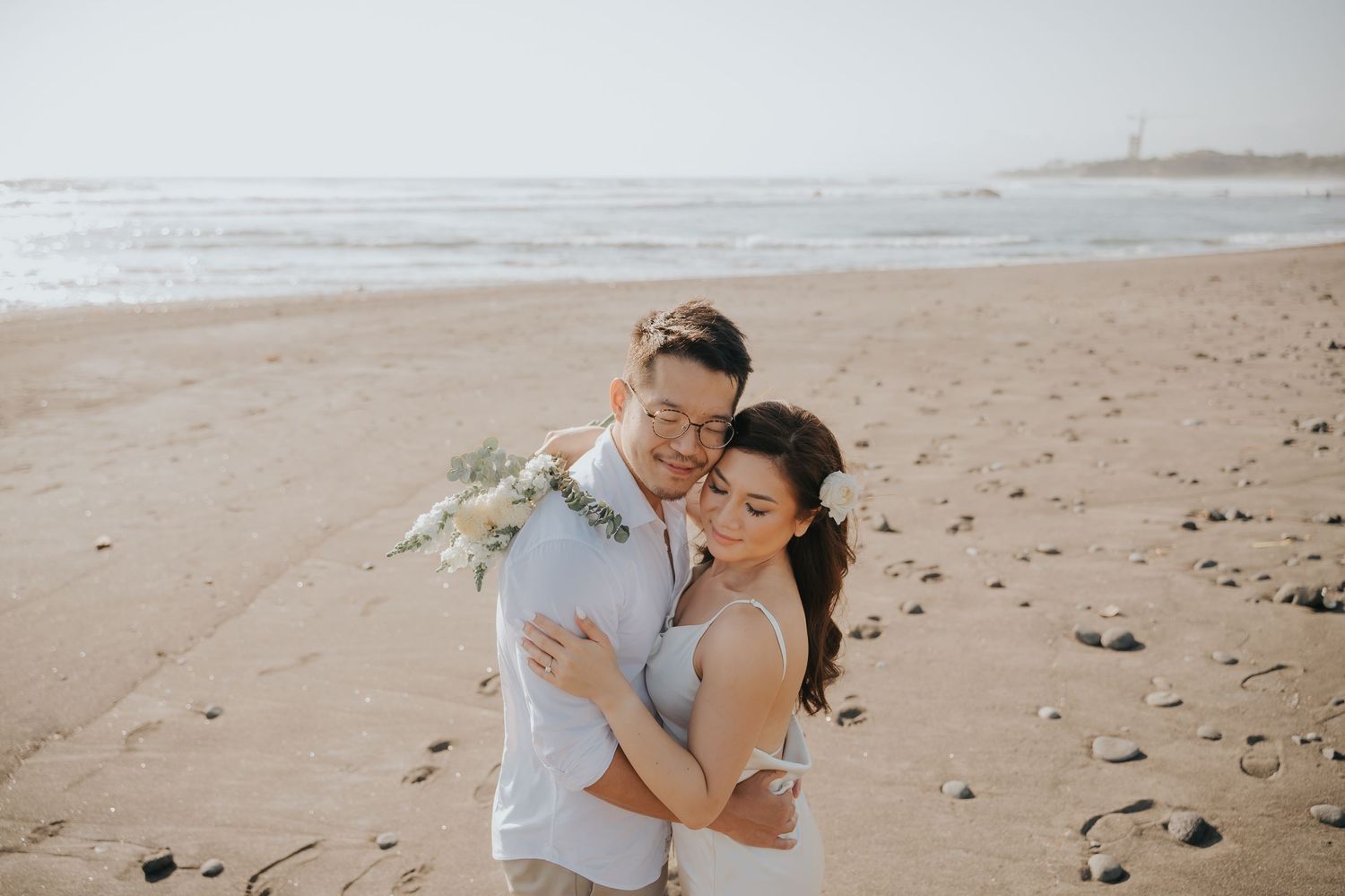 Couple embracing on sandy beach during golden hour, woman in white dress holding bouquet, romantic seaside wedding photo.