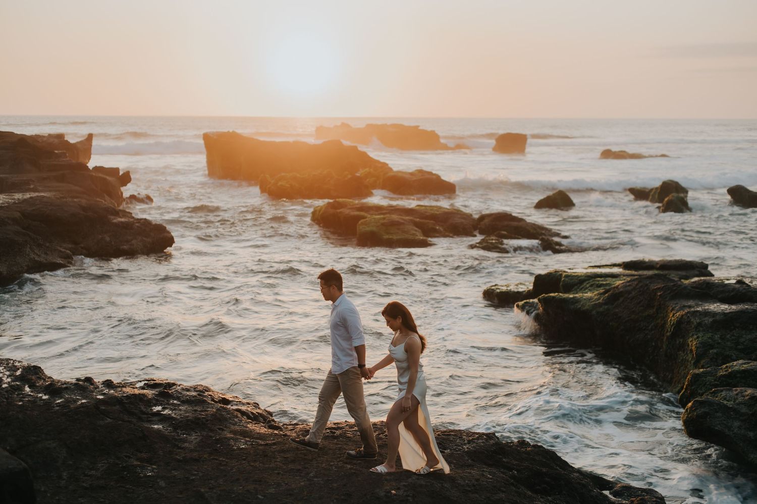Couple holding hands walking on rocky coastline at golden sunset with ocean waves crashing on rocks behind them.