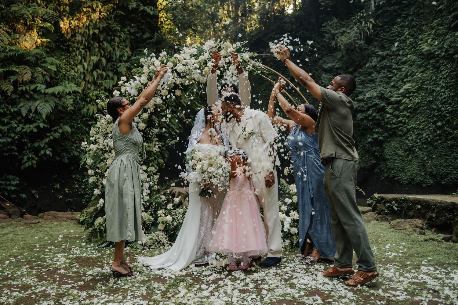 Joyful wedding party tossing white flower petals over bride and groom beneath floral arch outdoors.