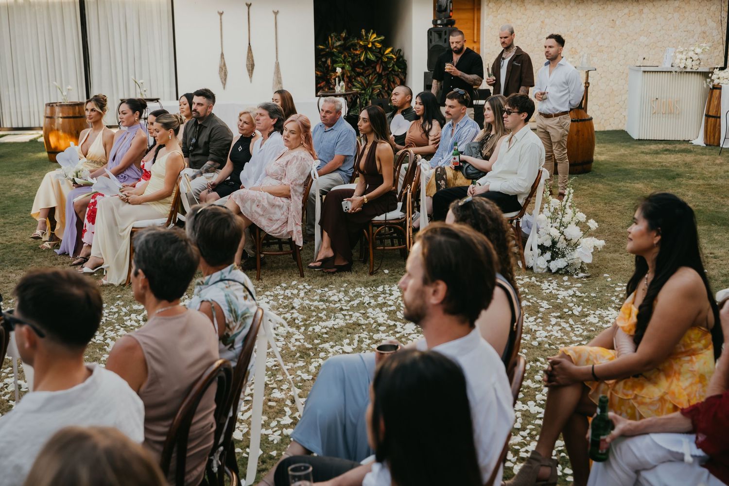 Outdoor wedding ceremony with guests seated on wooden chairs along a white petal-lined aisle.
