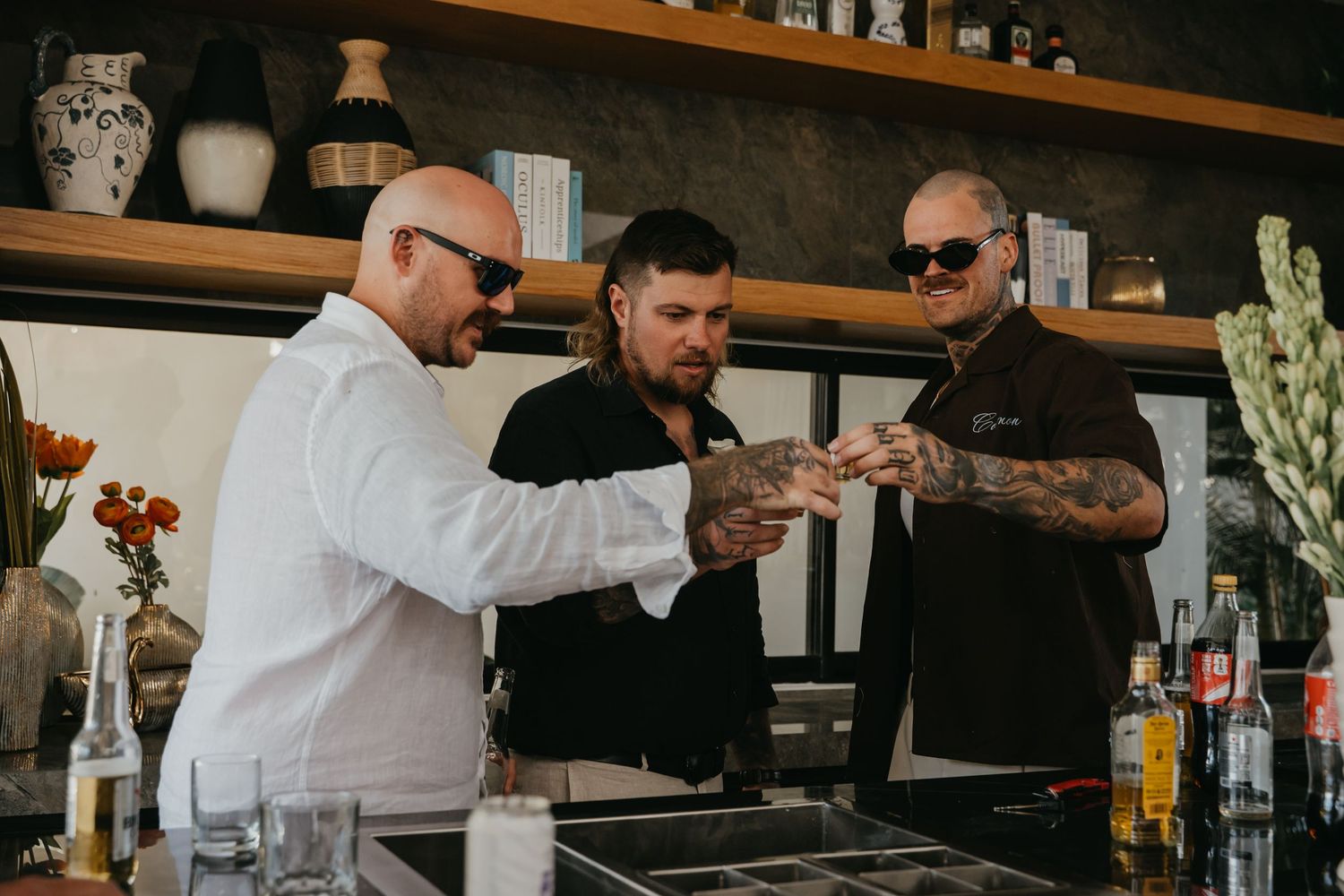 Three tattooed bartenders examining cocktail ingredients at a stylish bar with bottles and glassware.