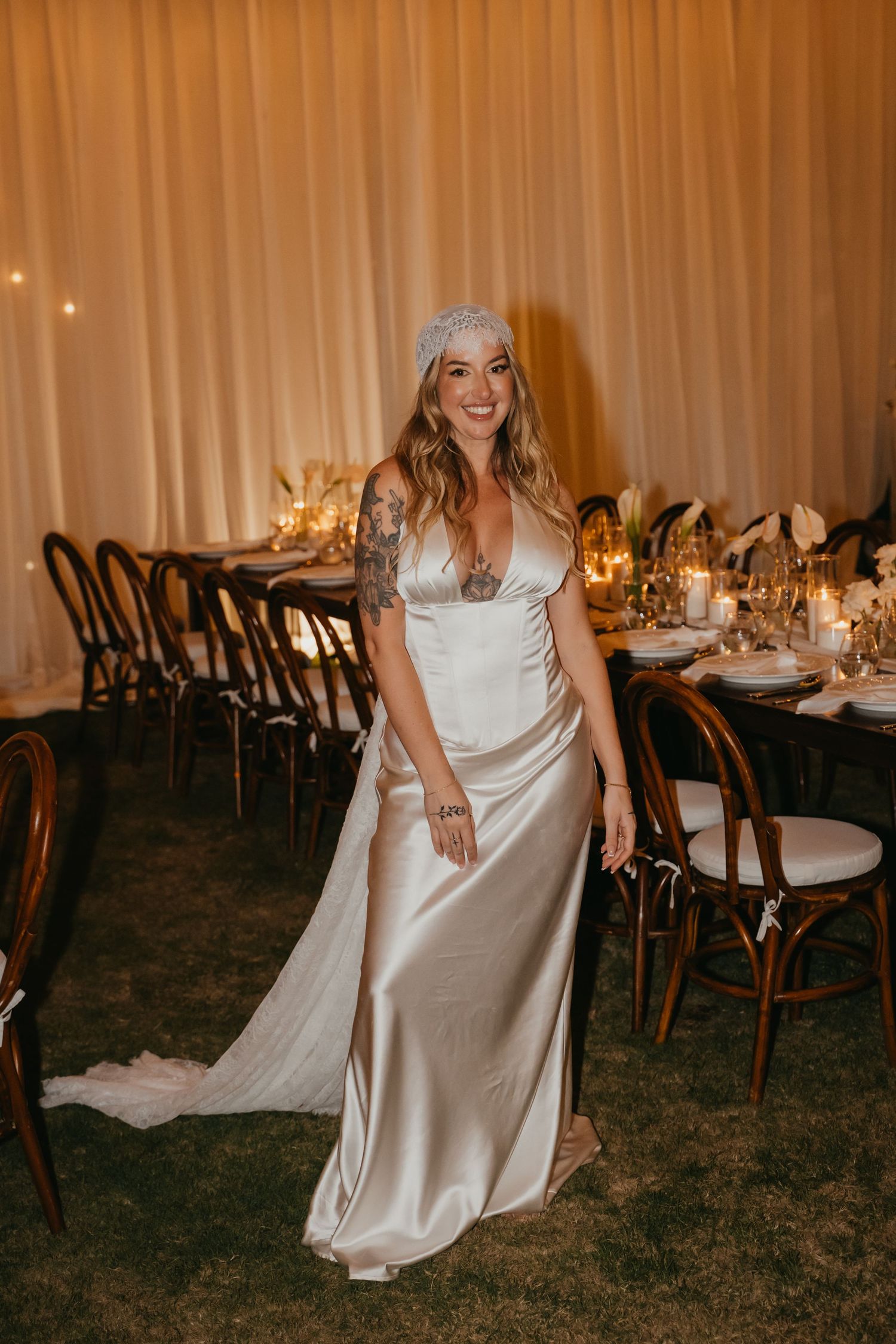 Tattooed bride in satin wedding gown and lace headpiece smiles in reception tent.