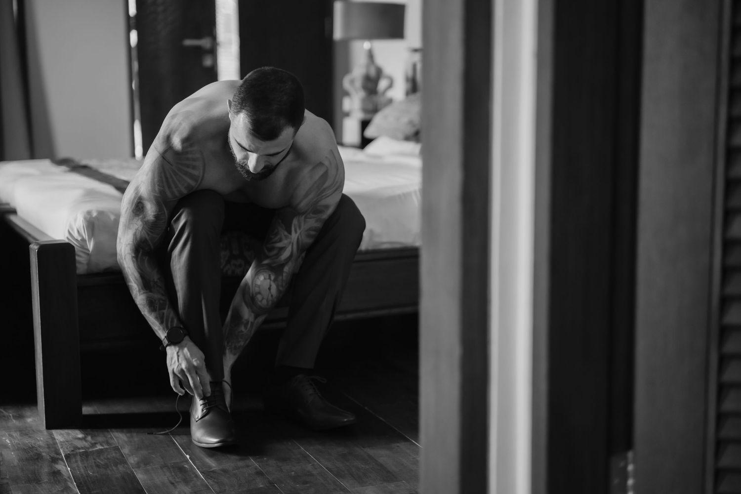 Black and white photo of a muscular tattooed man tying his dress shoes while sitting in a bedroom.
