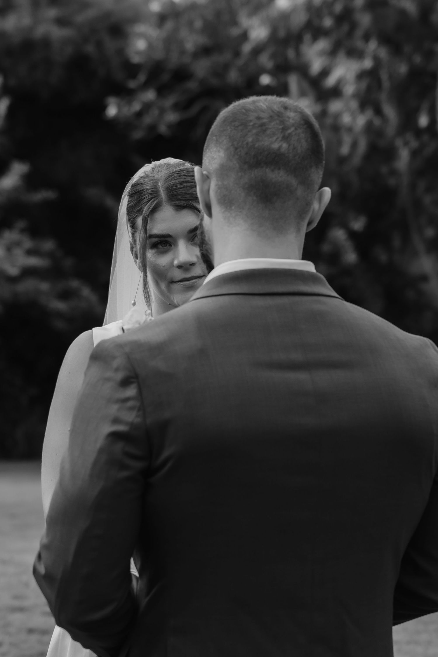 Black and white portrait of bride in veil gazing at groom during ceremony.
