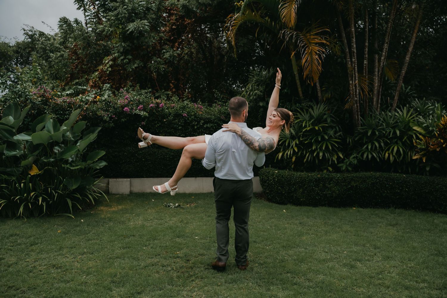 Groom lifts bride joyfully in the air during outdoor wedding celebration in tropical garden.