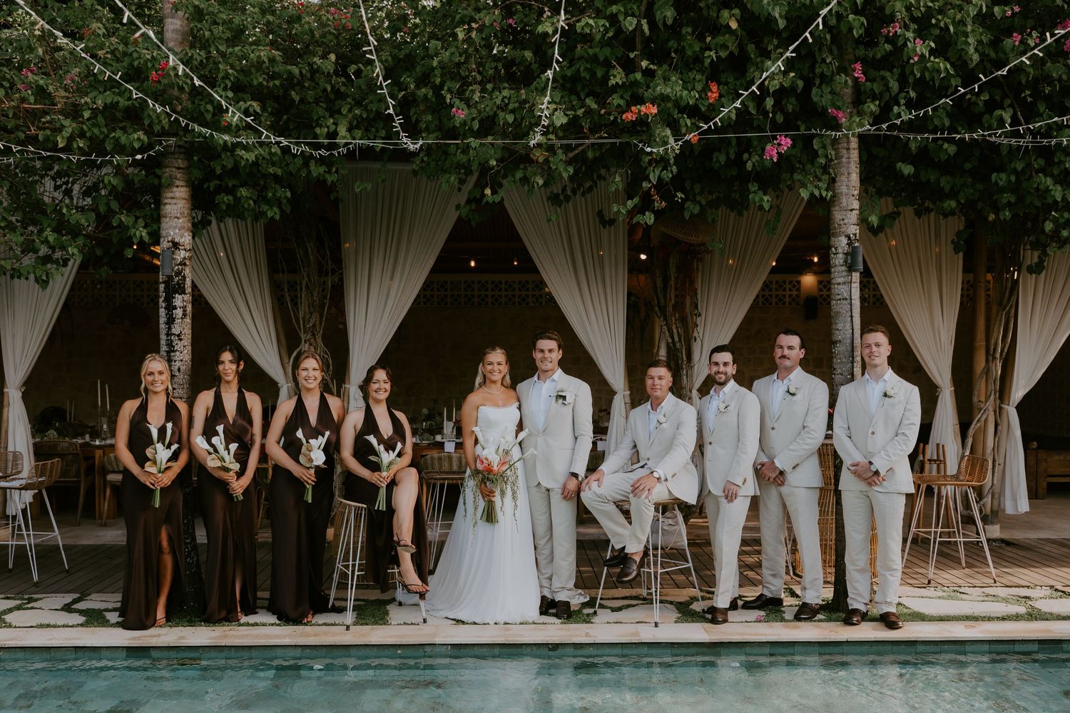 Wedding party poses poolside with bridesmaids in dark gowns and groomsmen in white suits under string lights.