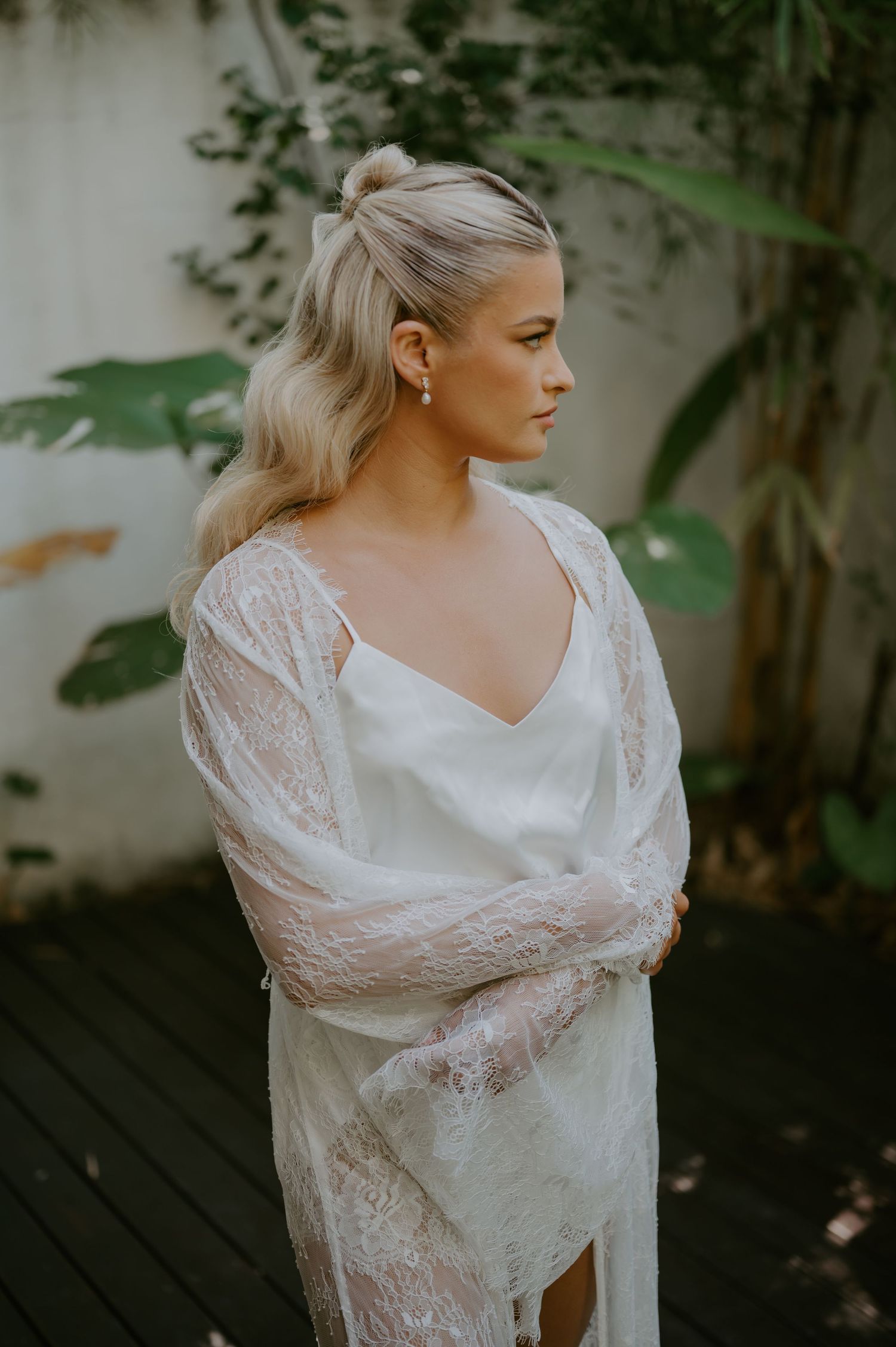 Elegant blonde bride in lace robe with half-up hairstyle and pearl earrings poses outdoors against tropical garden wall.