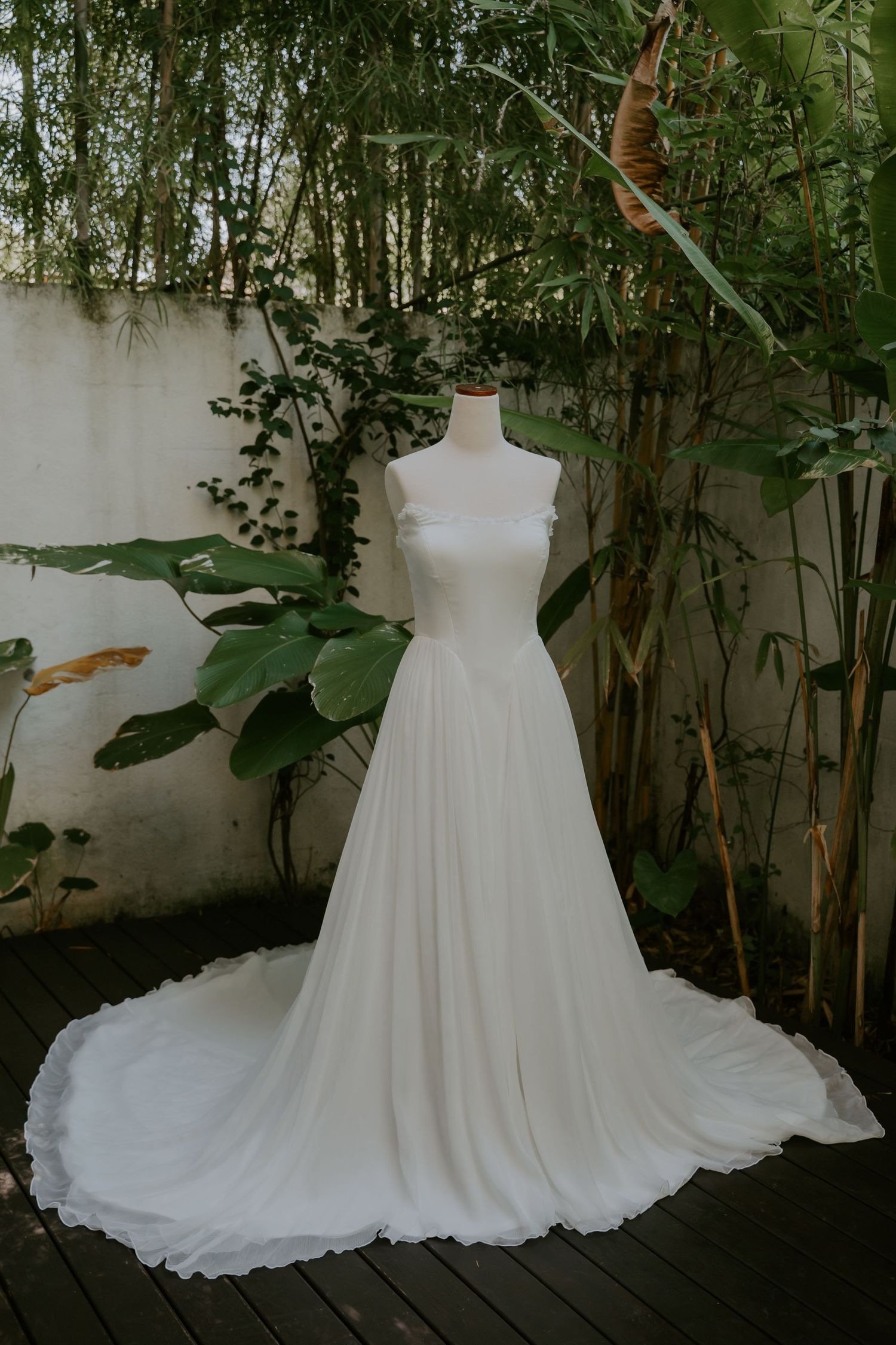 Elegant white wedding gown with flowing train displayed on a mannequin in a lush tropical garden setting.
