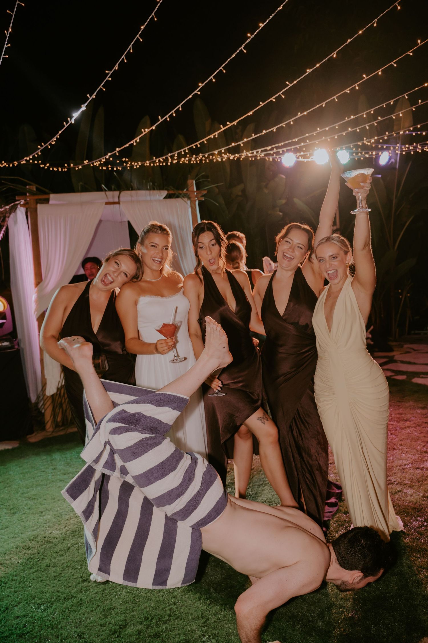 Bride poses with bridesmaids in dark gowns raising glasses under string lights at outdoor wedding reception.