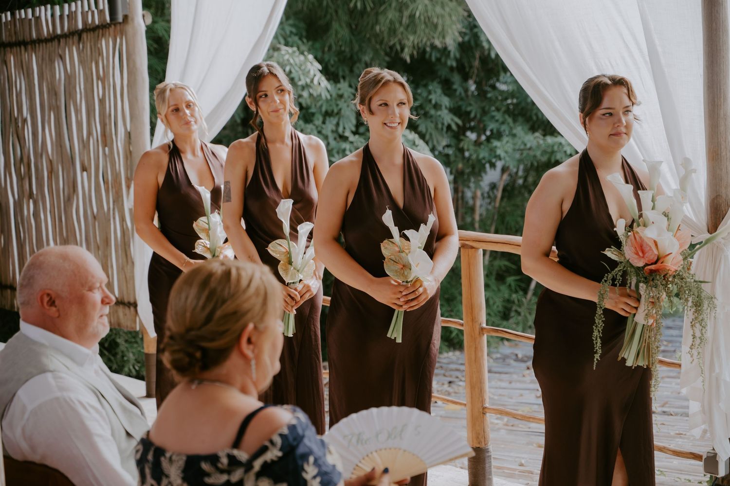 Four bridesmaids in dark brown multiway dresses hold white bouquets while standing at the wedding ceremony.