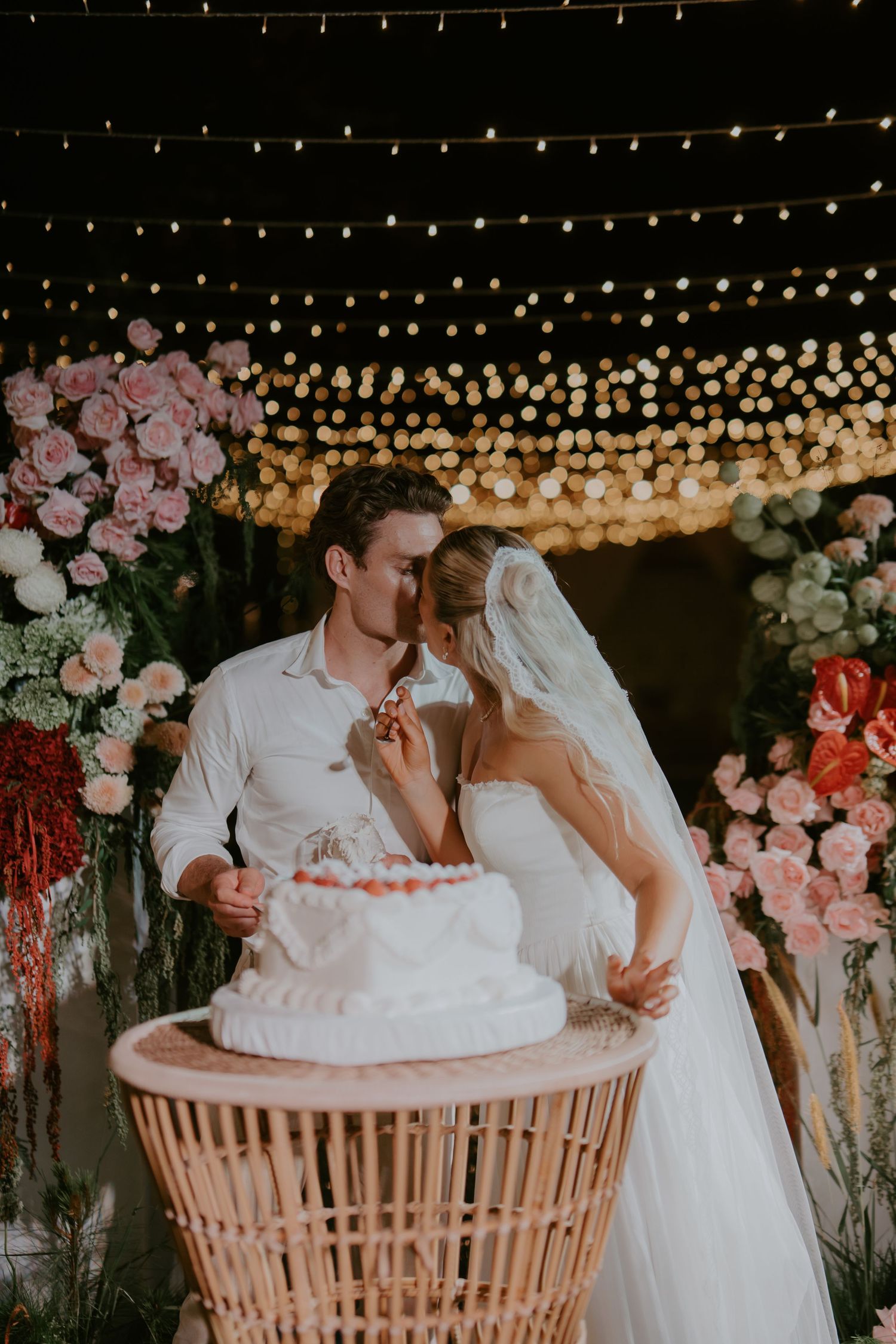 Newlyweds kiss while cutting white wedding cake surrounded by roses and string lights.