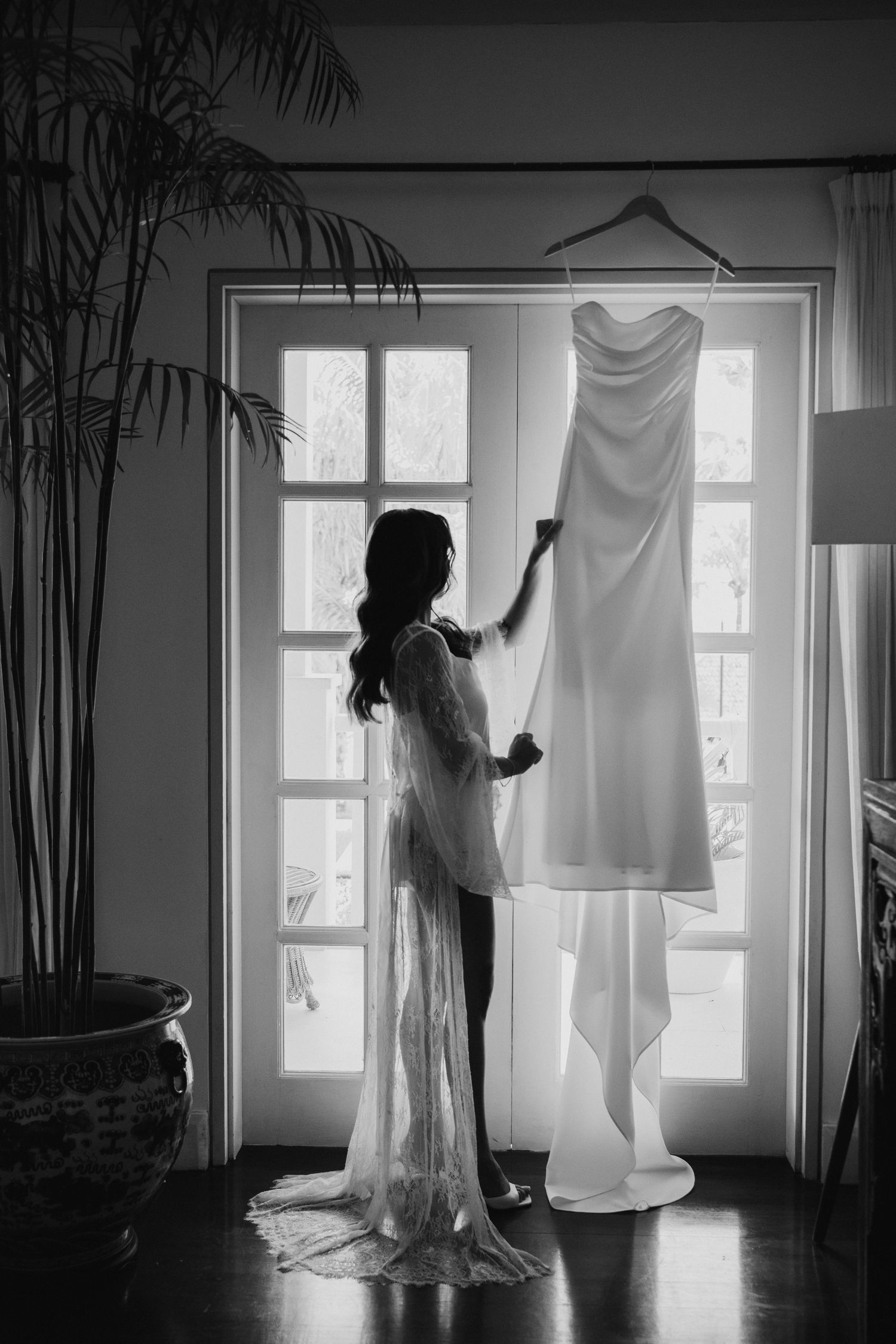 Bride in lace robe admires her wedding gown hanging by a sunlit window in black and white photo.
