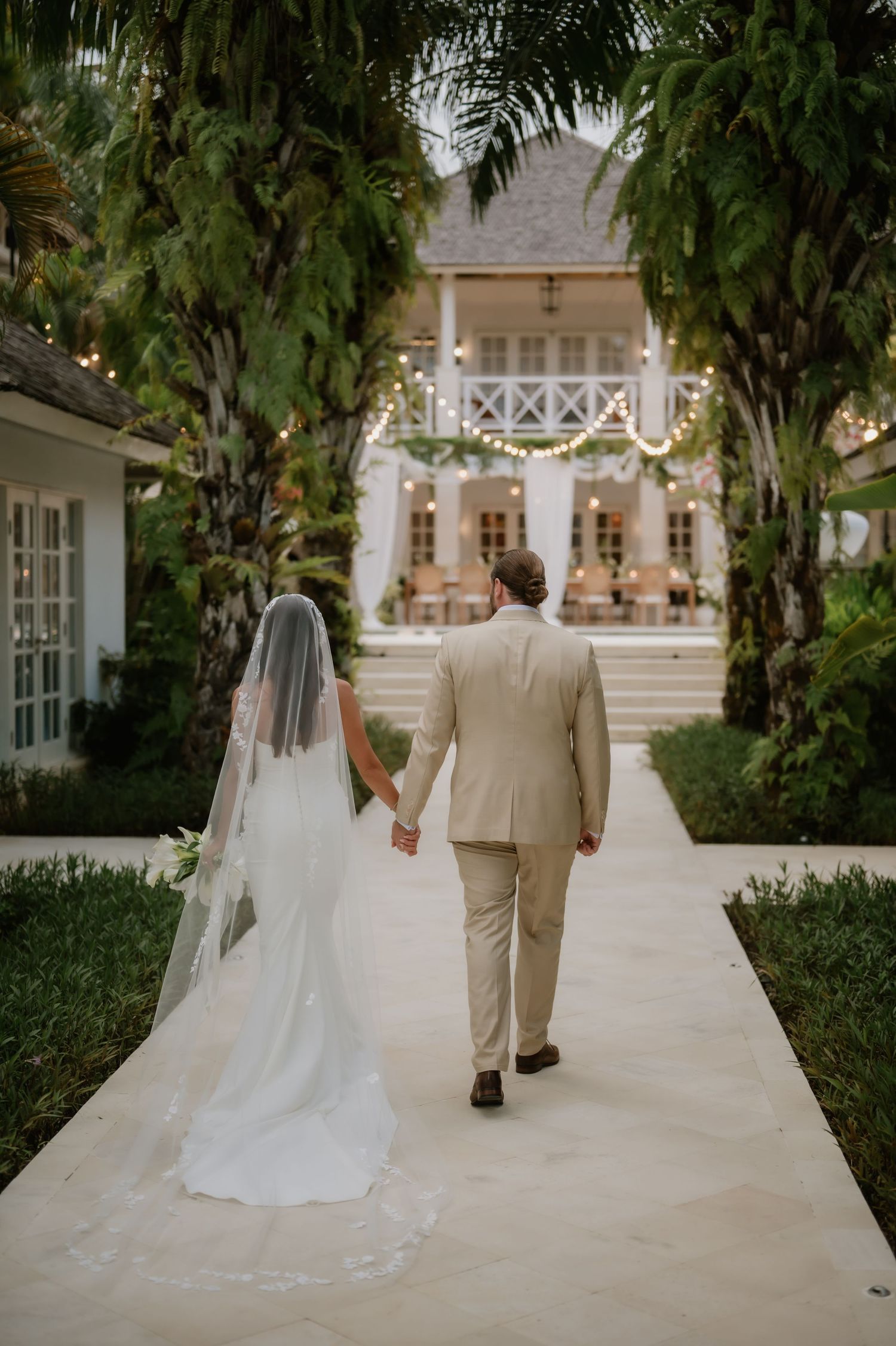 Bride and groom walk hand-in-hand toward a tropical wedding venue adorned with string lights and lush greenery.
