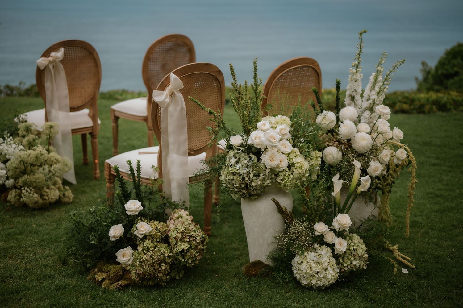 Elegant outdoor wedding ceremony setup with white floral arrangements, gold chairs, and ocean backdrop on lush green lawn.