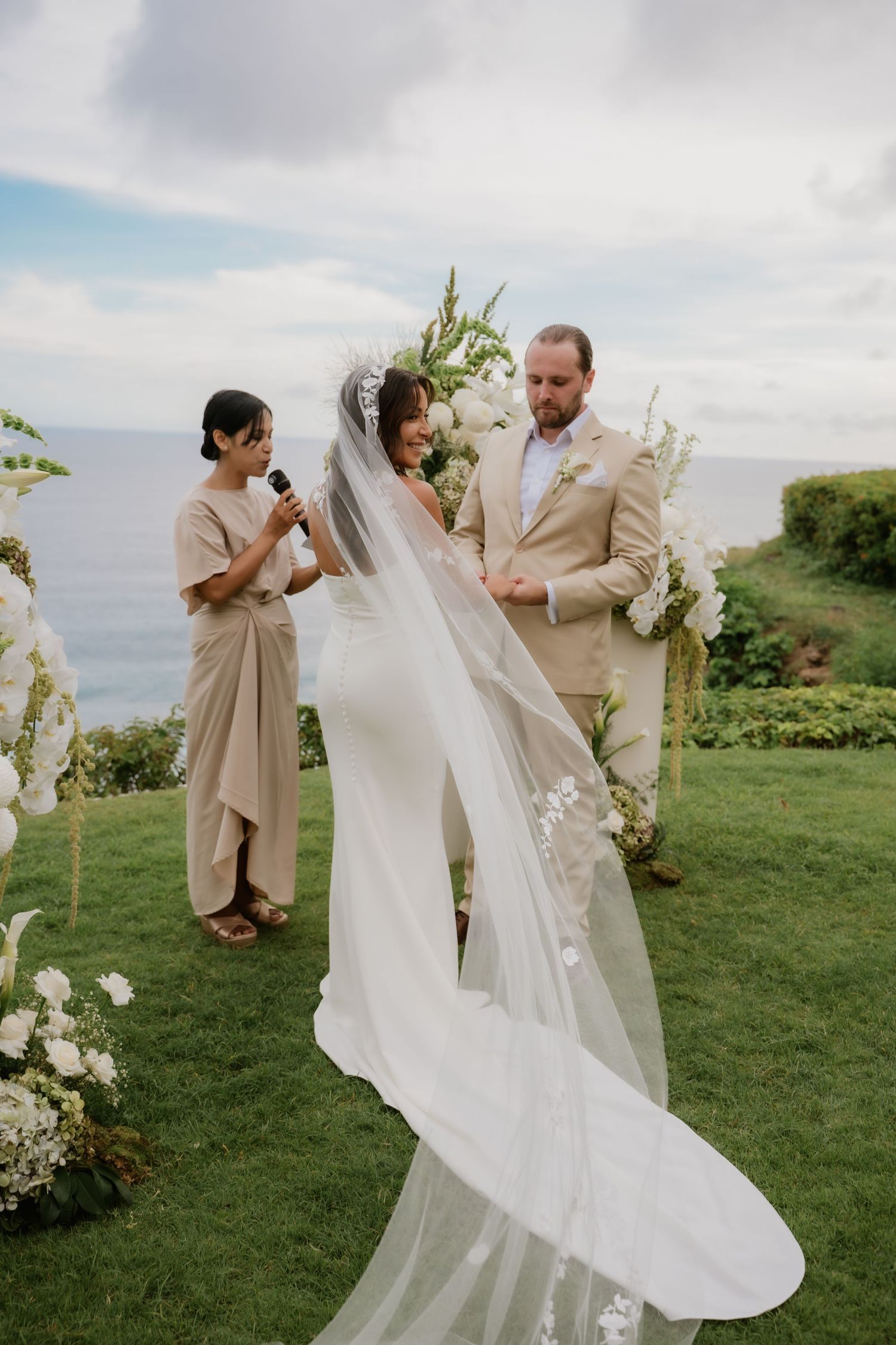 Bride and groom exchange vows at an outdoor cliffside wedding ceremony with white floral arch and ocean backdrop.