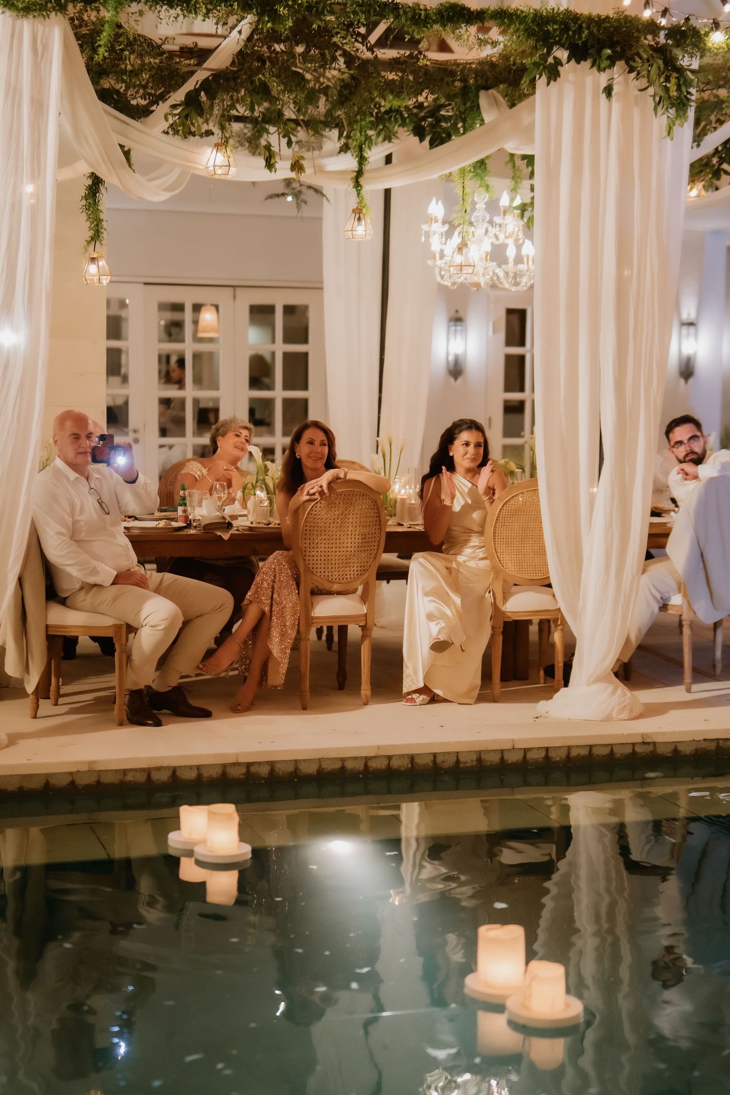 Wedding guests dine at elegantly decorated poolside reception with floating candles and draping.