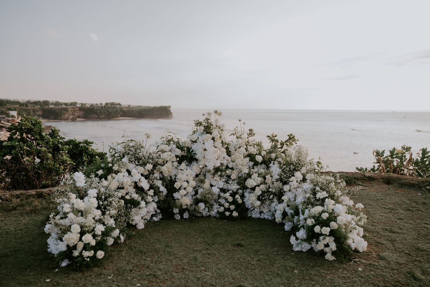 Stunning white floral arch wedding ceremony backdrop set against ocean cliff view.