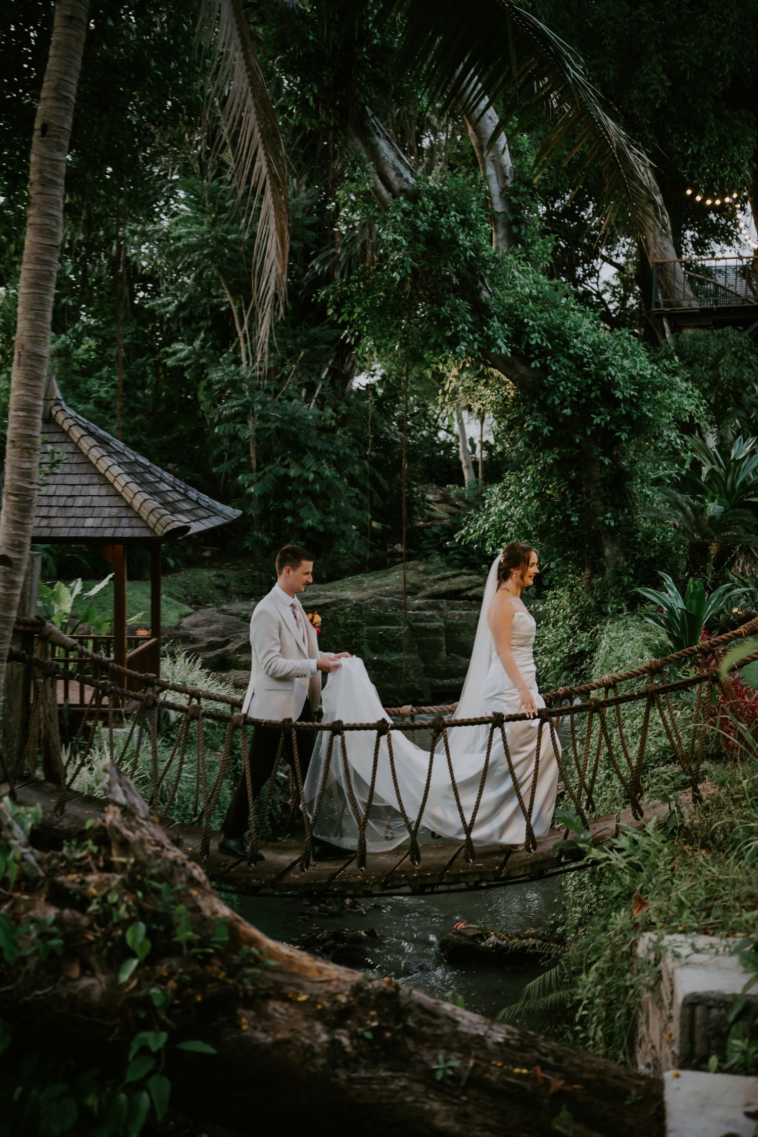 Bride and groom crossing rustic rope bridge through lush tropical jungle on their wedding day.