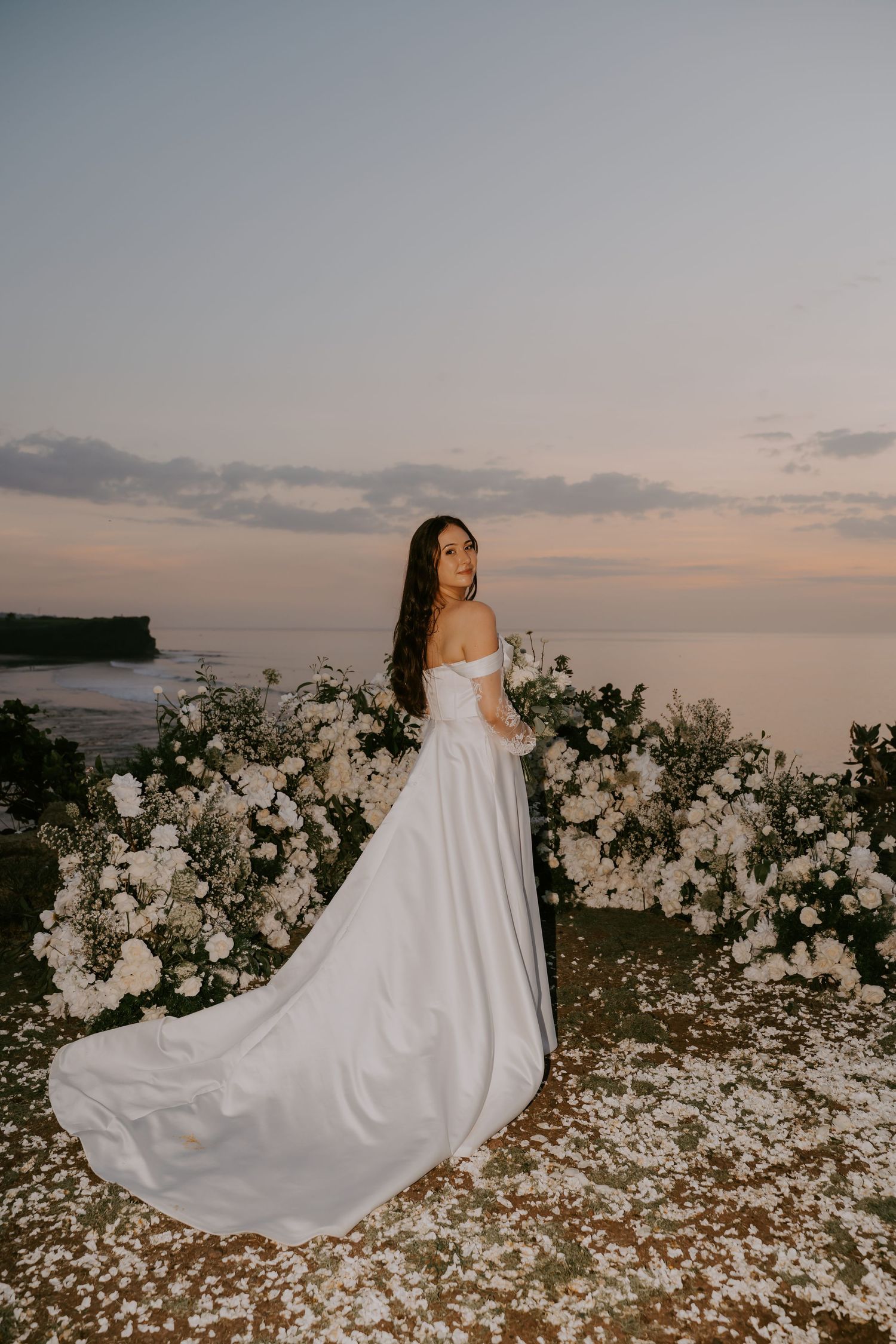 Bride in off-shoulder white gown with train poses before white floral arch at sunset ocean wedding.