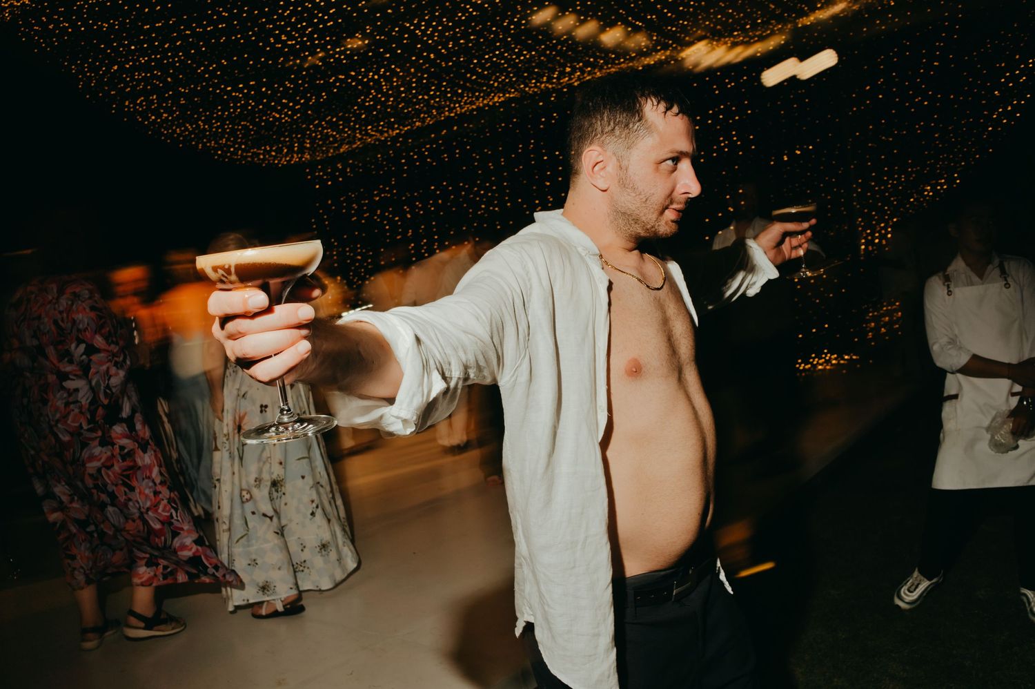 Wedding guest dancing shirtless while holding a cocktail under a canopy of fairy lights.