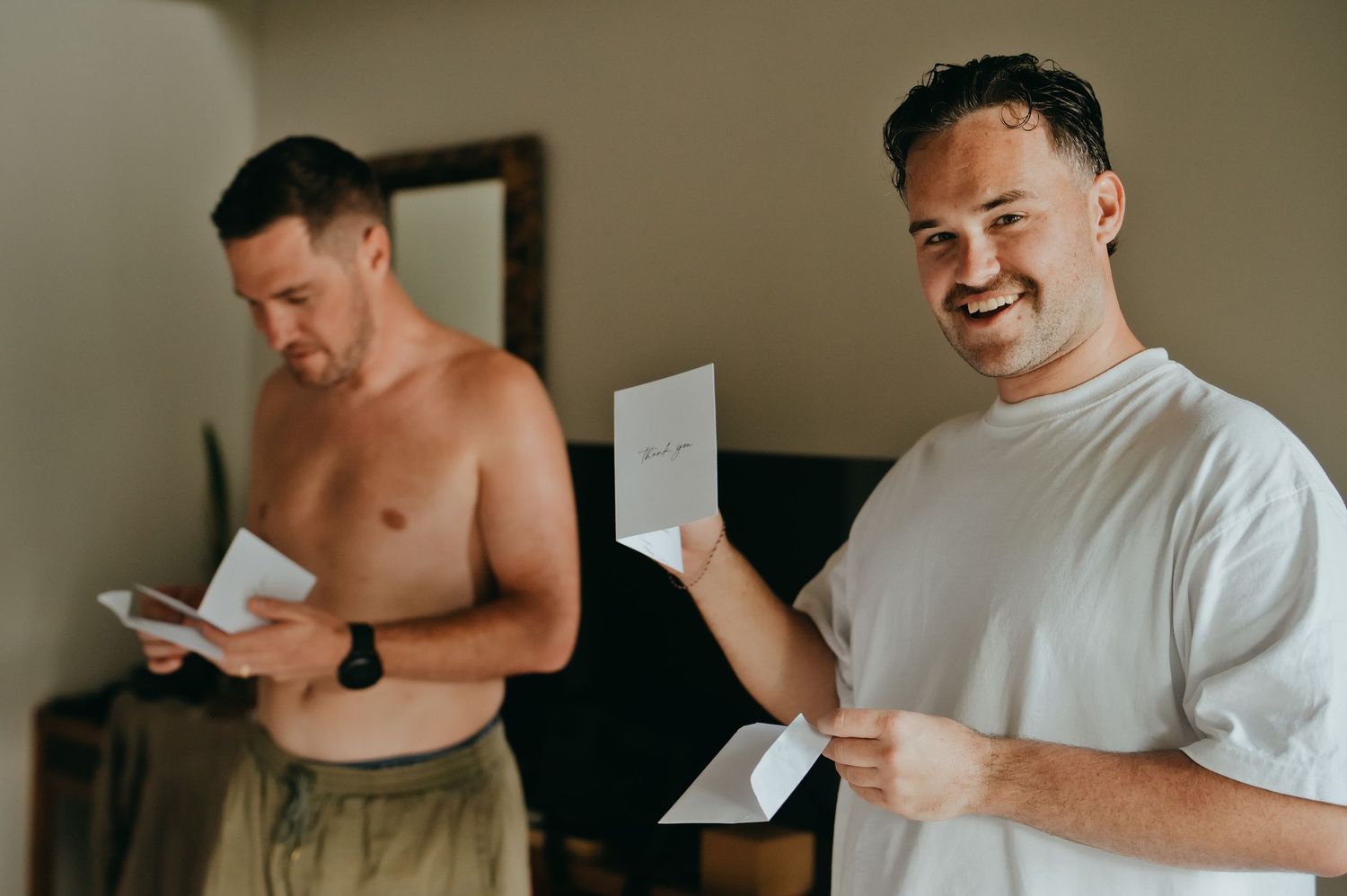 Two groomsmen smiling while reading heartfelt notes during wedding morning preparations indoors.