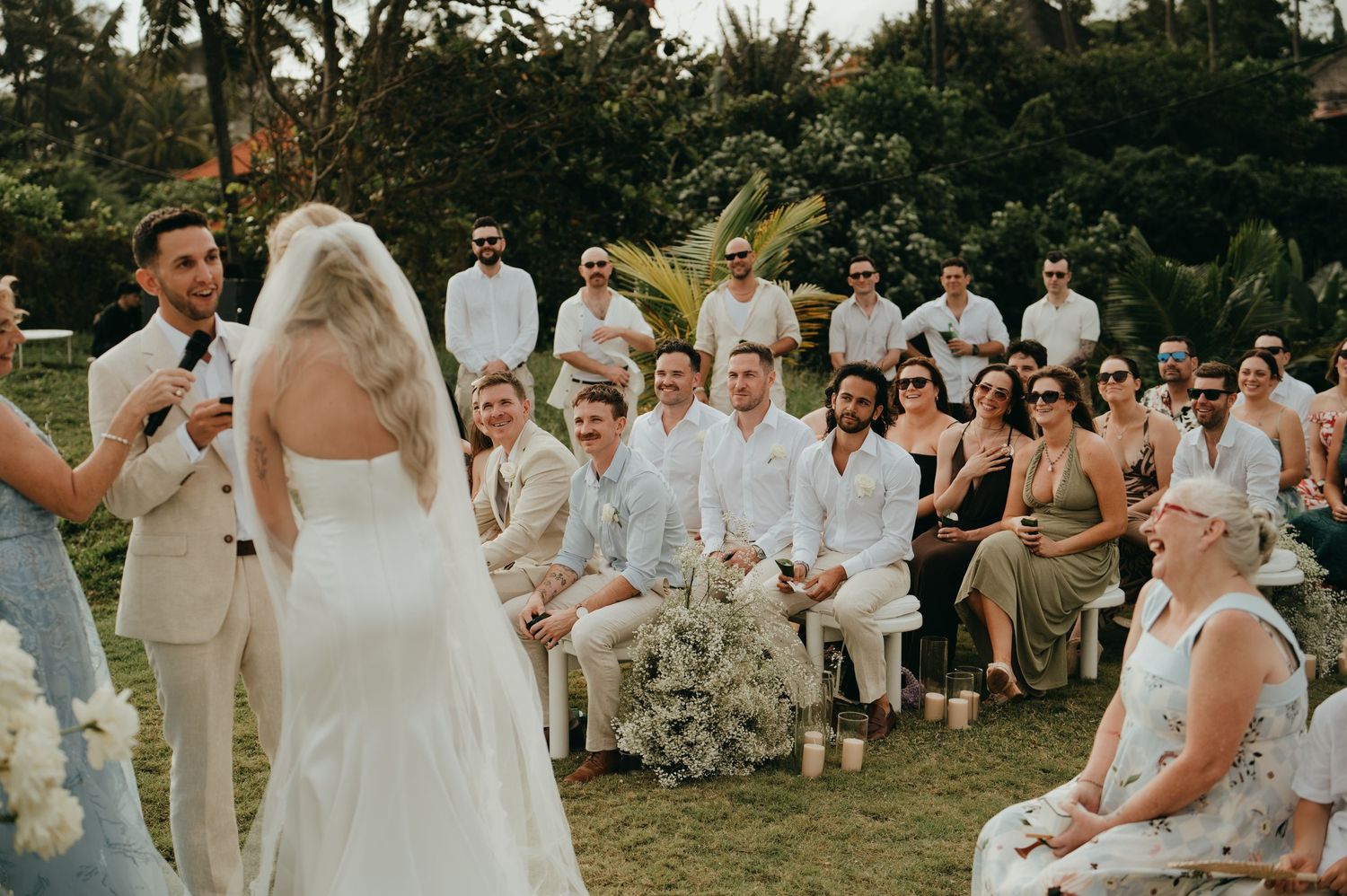 Bride in white gown faces groom during outdoor tropical wedding ceremony with seated guests.