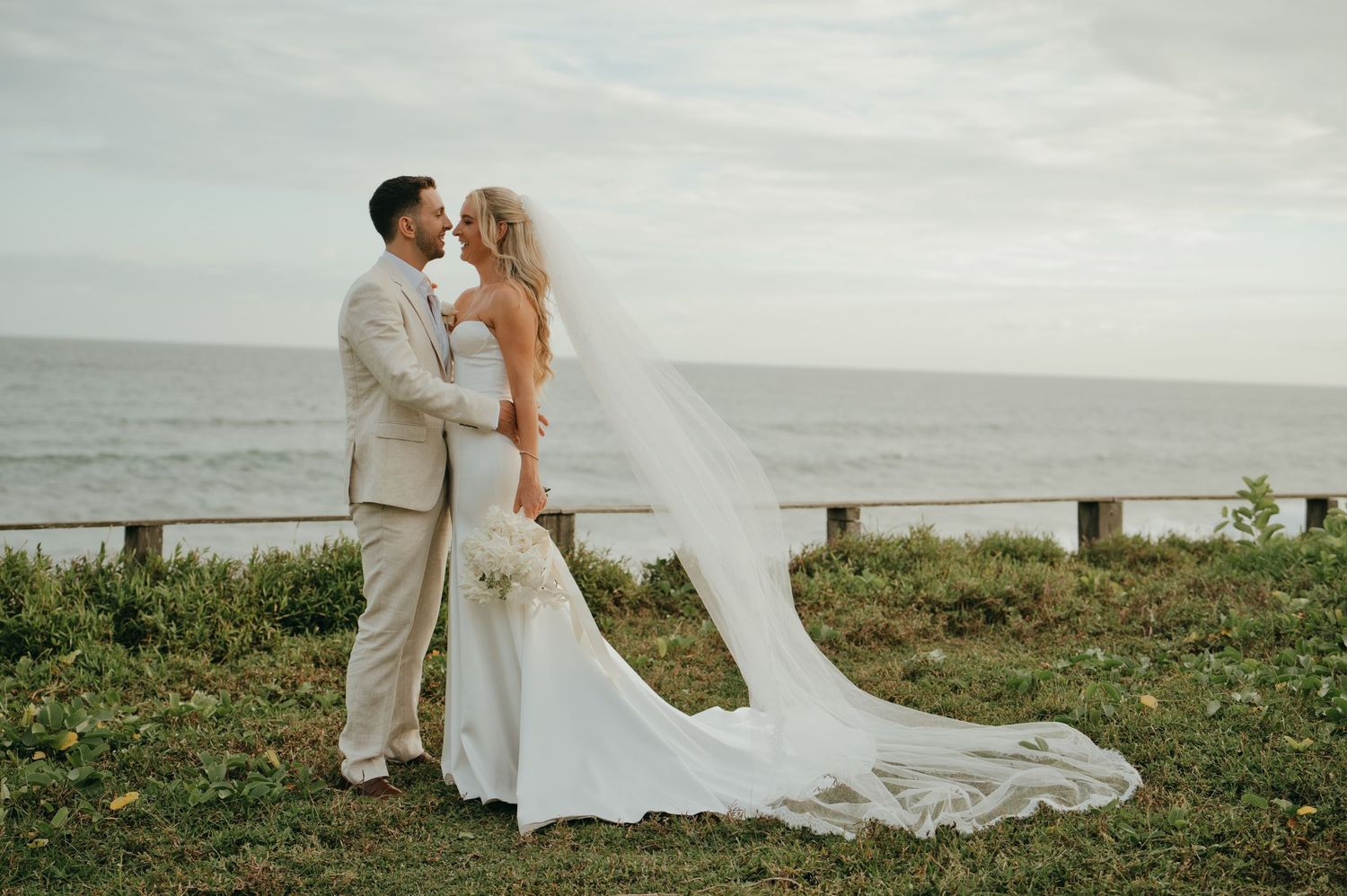 Bride and groom share a romantic kiss overlooking the ocean, with the bride's veil flowing in the breeze.