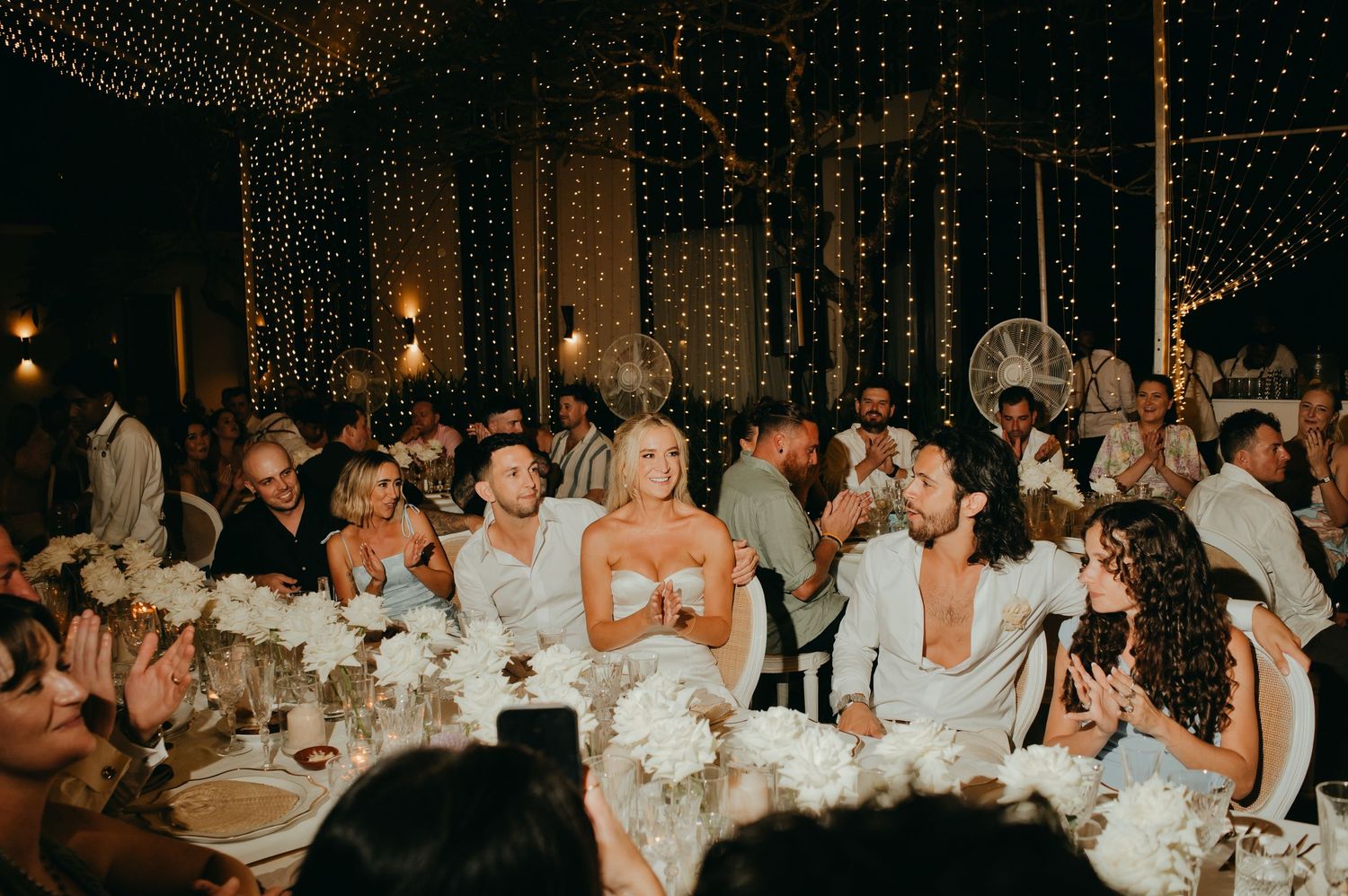 Bride and wedding guests laughing and celebrating at an elegant reception with white floral centerpieces.