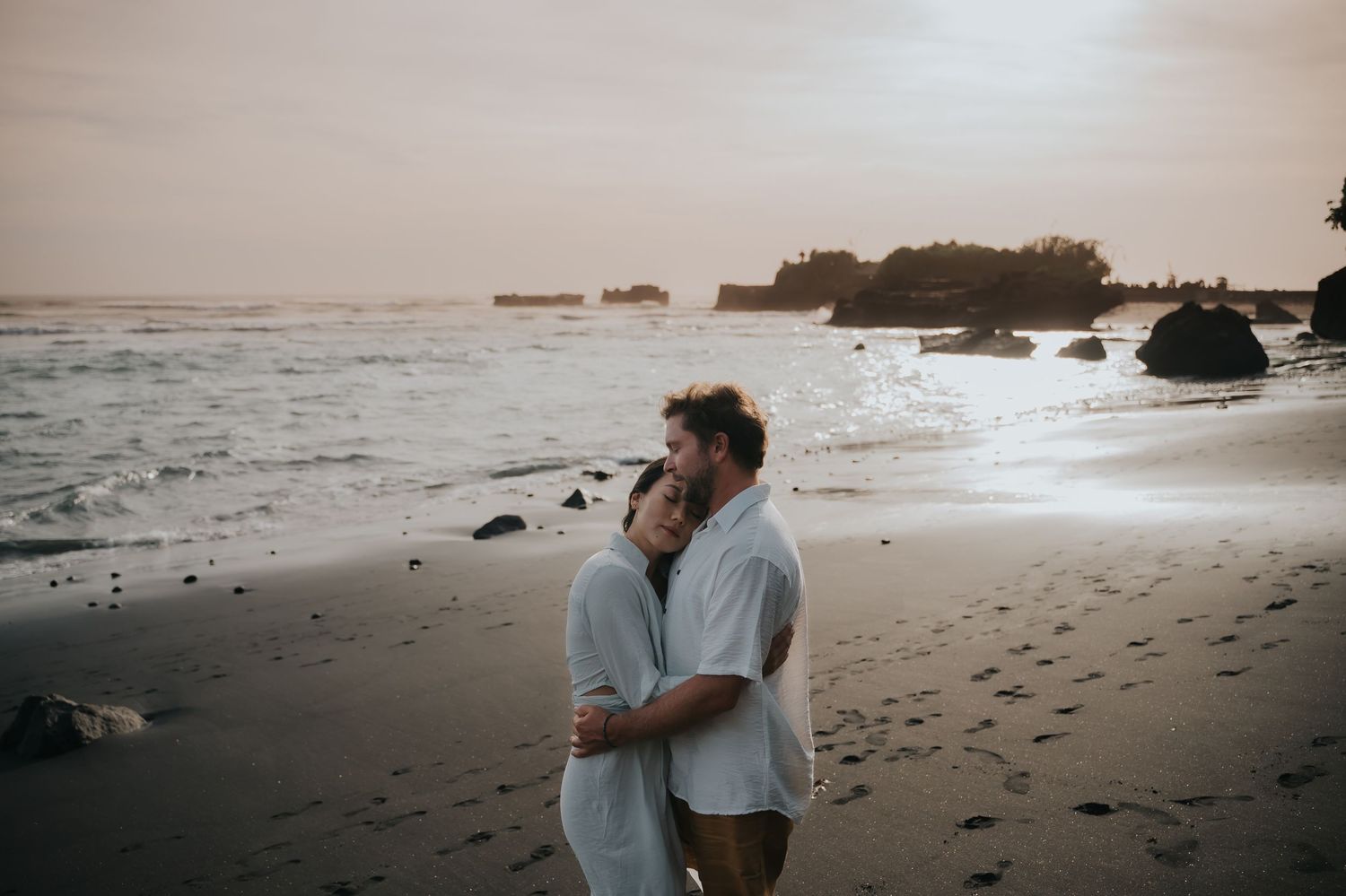 A couple embracing on a misty beach at sunset with waves and rocks in the background.