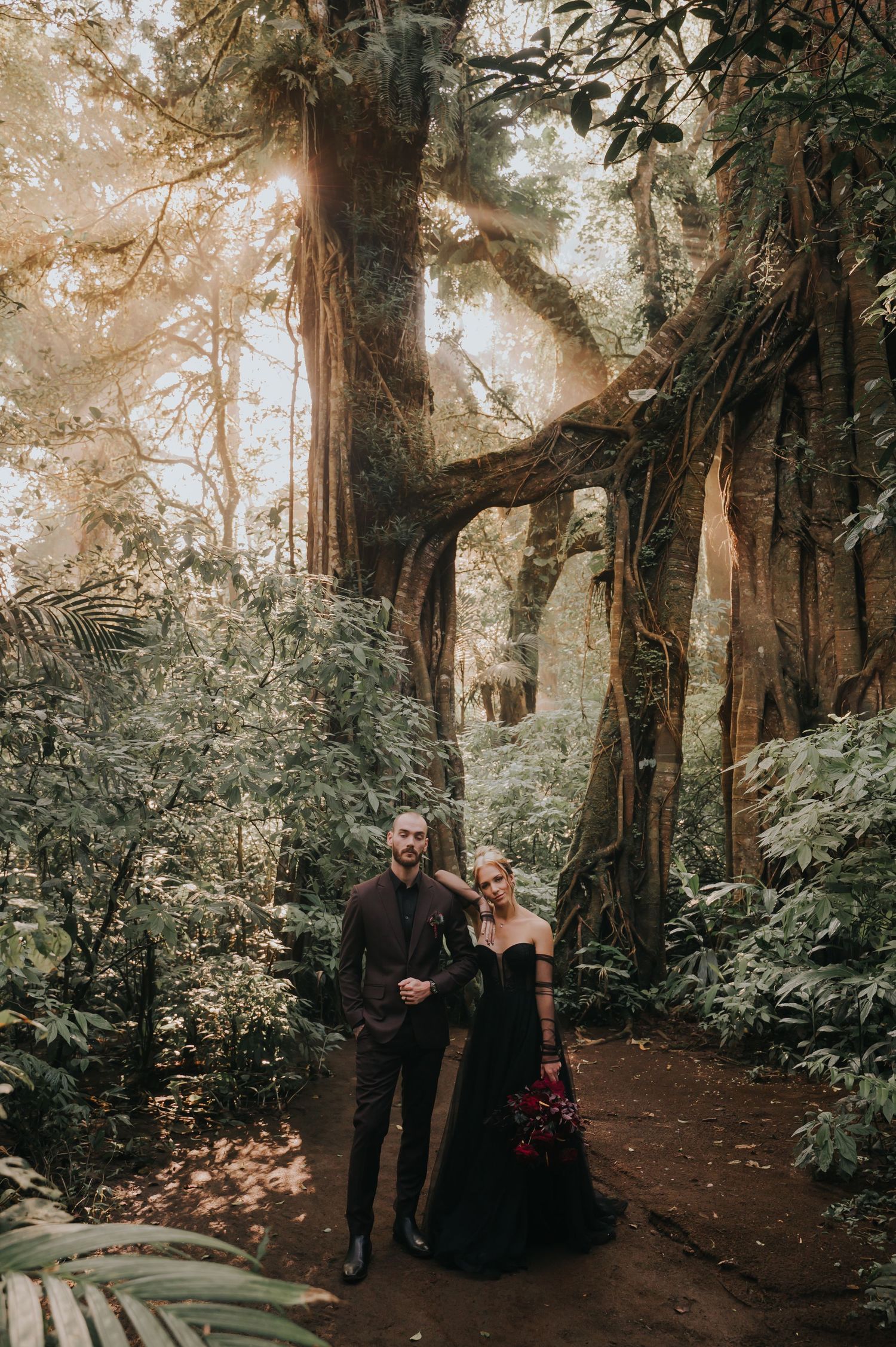 A couple poses on a forest path surrounded by towering redwood trees and lush greenery.