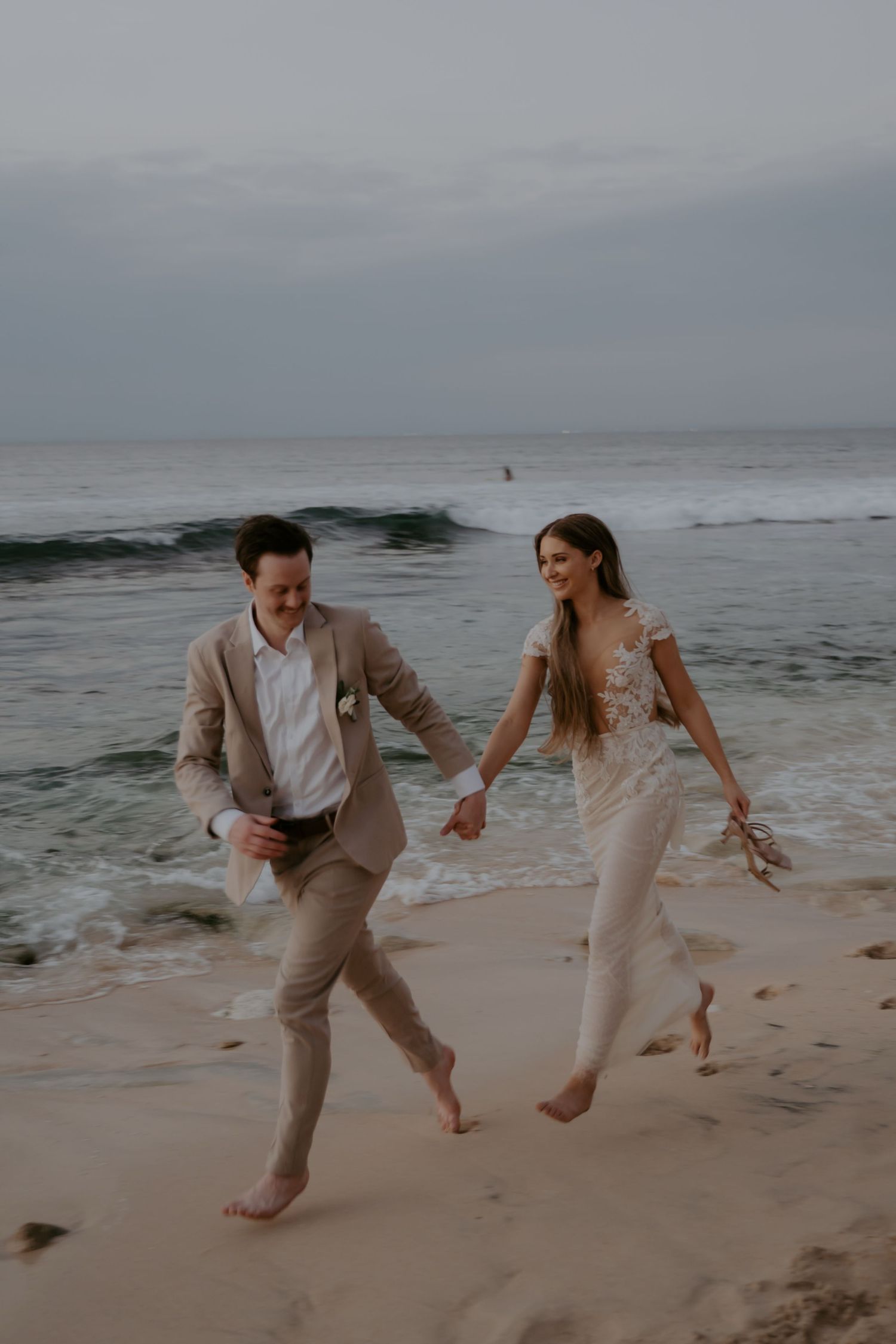 A couple in formal attire runs joyfully along a beach at sunset with waves crashing in the background.