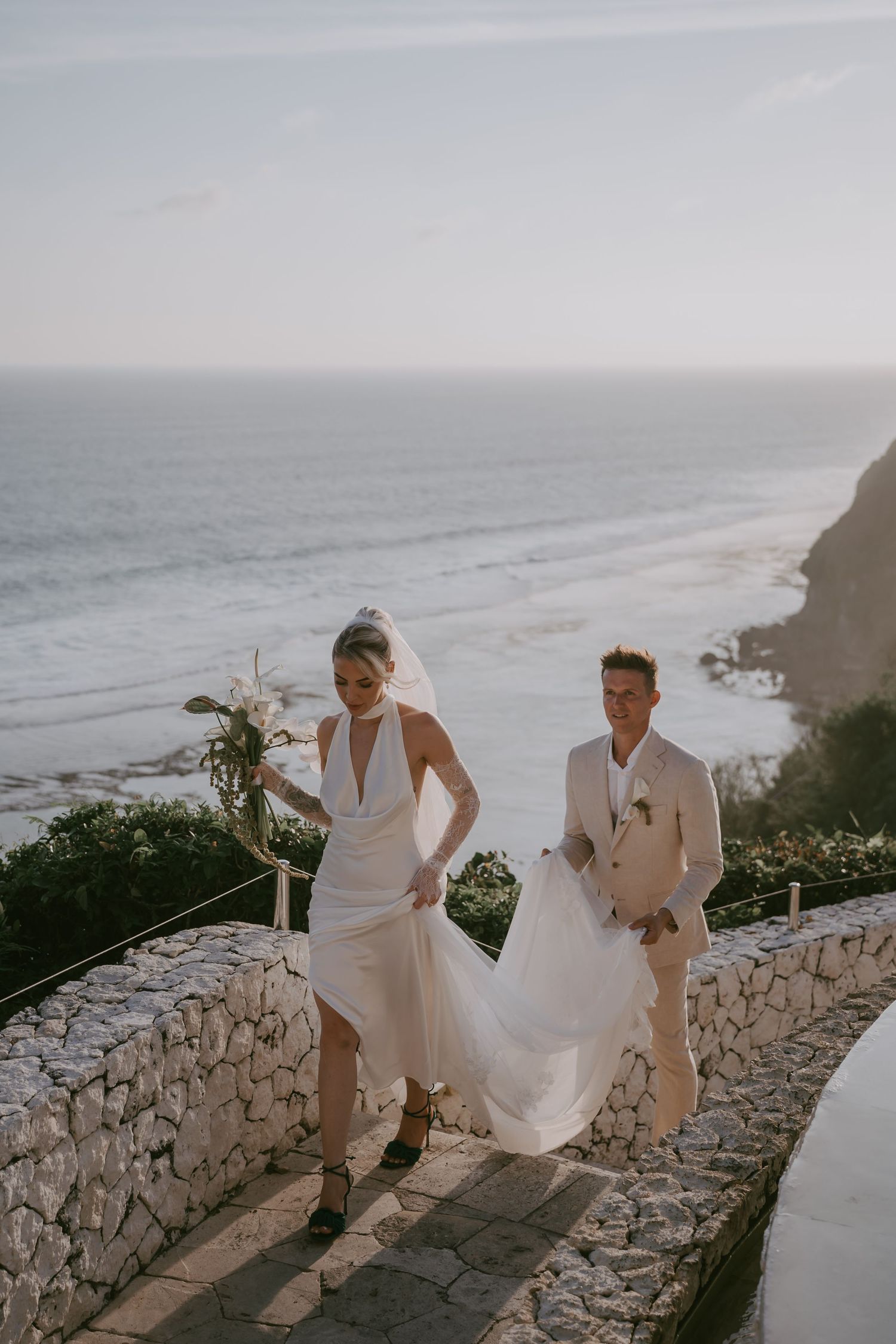 Couple in white attire walking along a scenic coastal stone path during sunset.