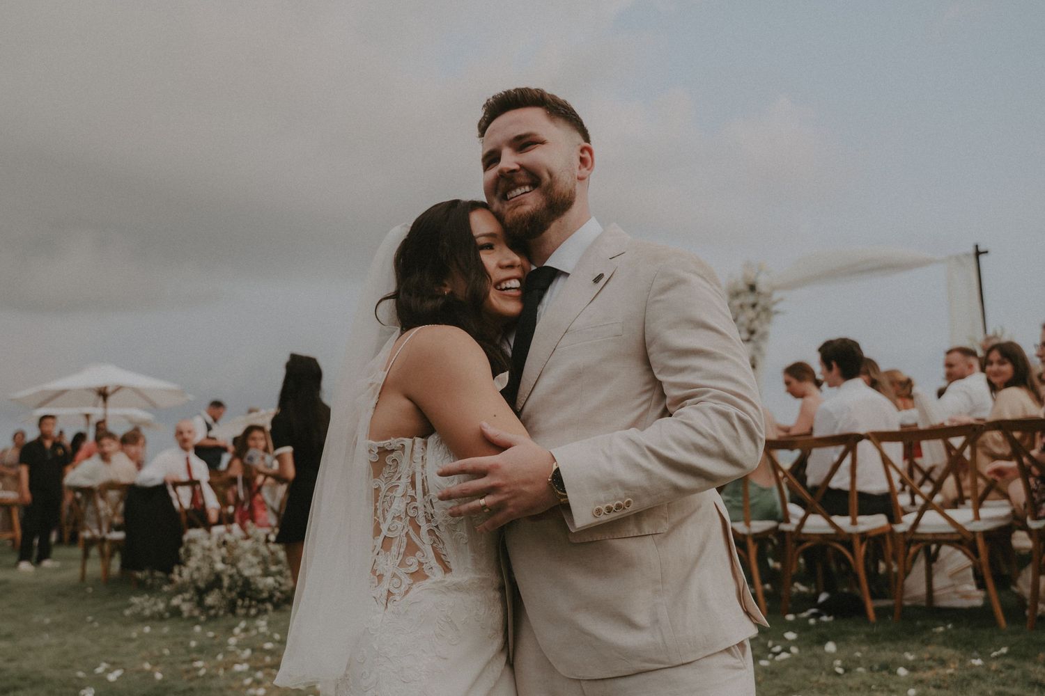 A couple shares a tender moment during an outdoor wedding ceremony with guests seated in the background.